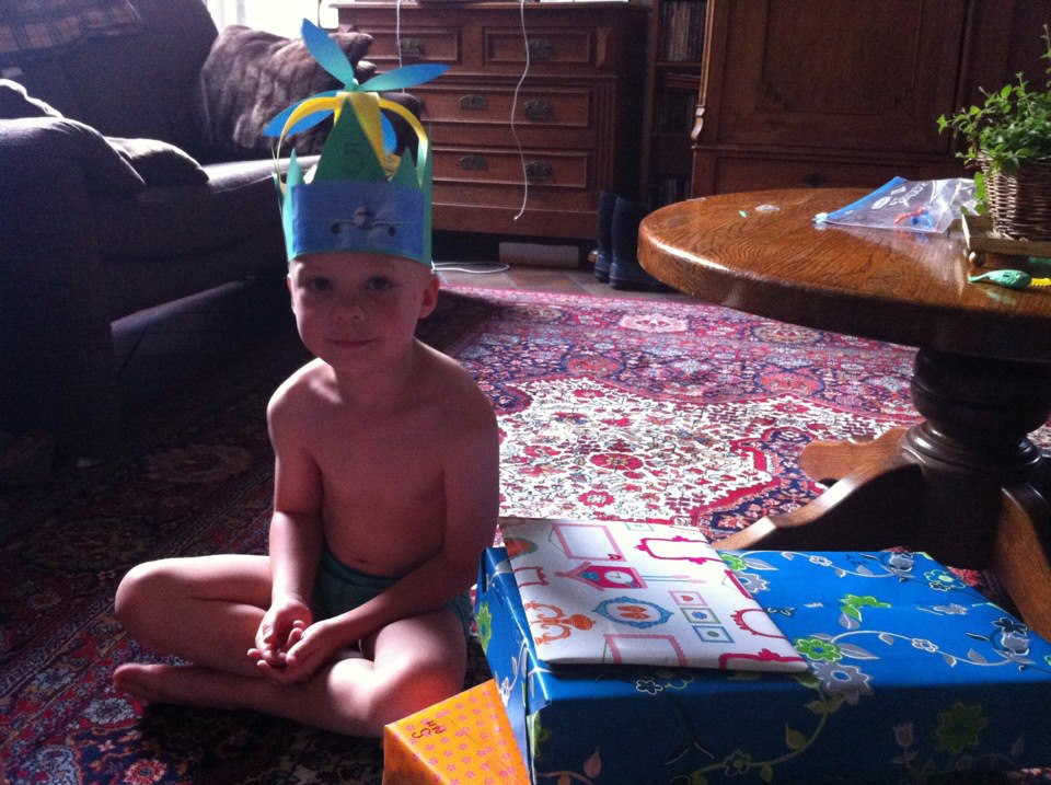 A young child wearing a handmade paper crown sits on the floor next to wrapped birthday presents.