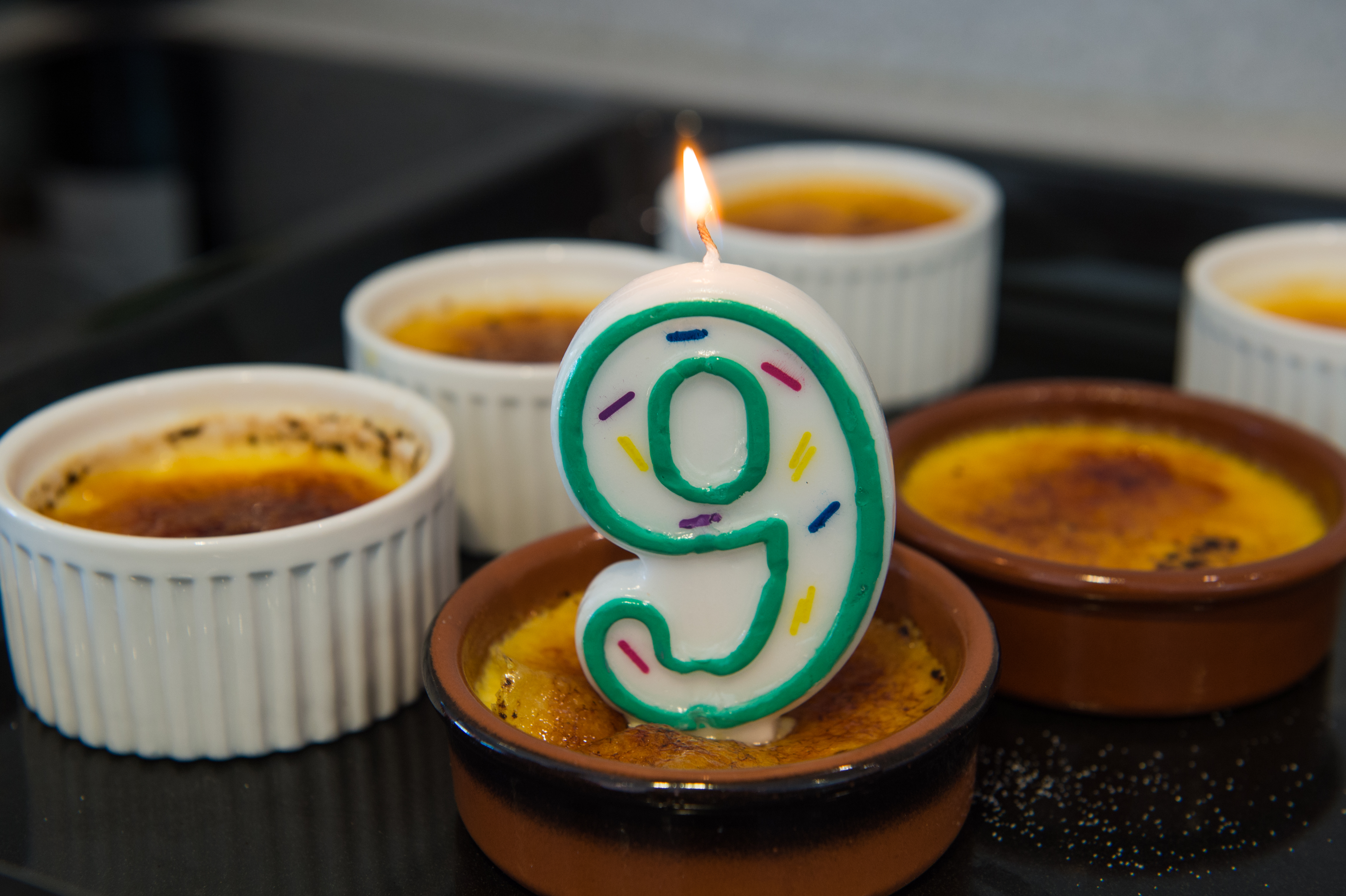 A lit number nine candle sits on crème brûlée, surrounded by more ramekins of the dessert for a birthday celebration.