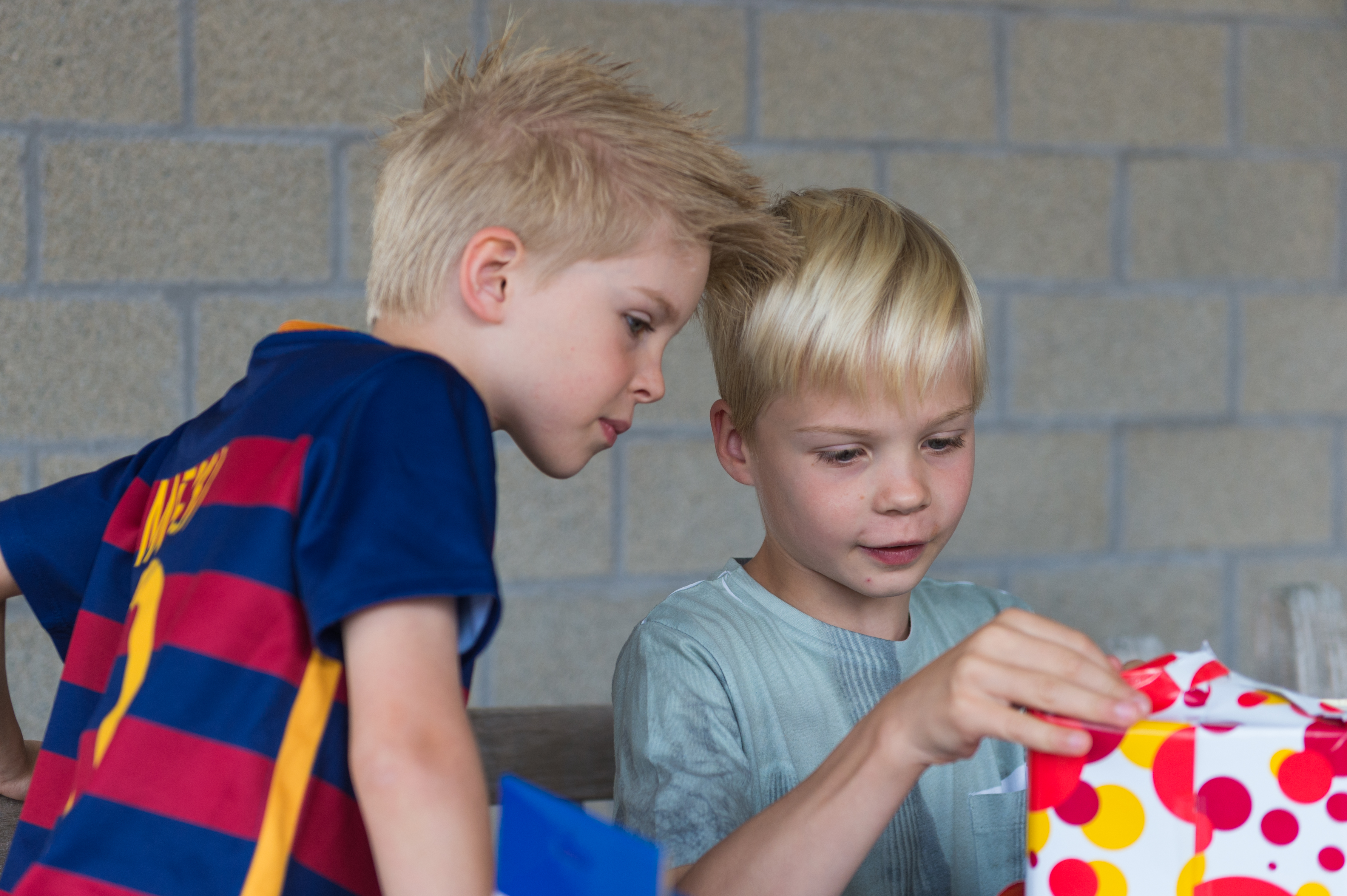 A young boy unwraps a colorful gift while another child watches closely beside him.