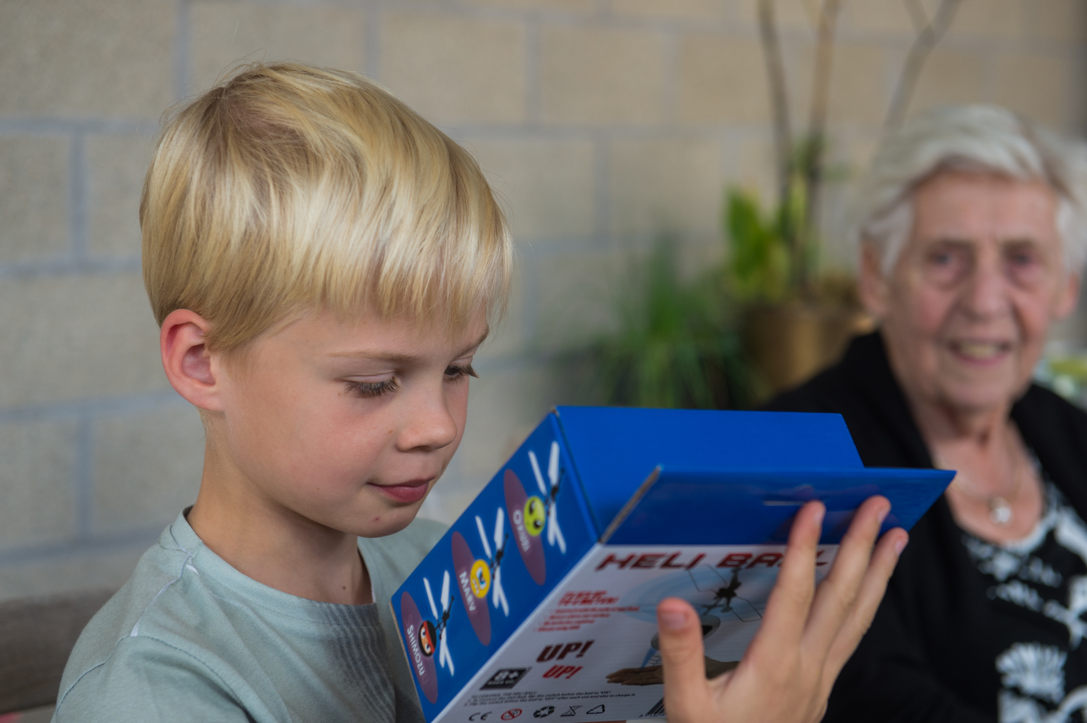 A young boy examines a blue toy helicopter box while an elderly woman watches and smiles in the background.