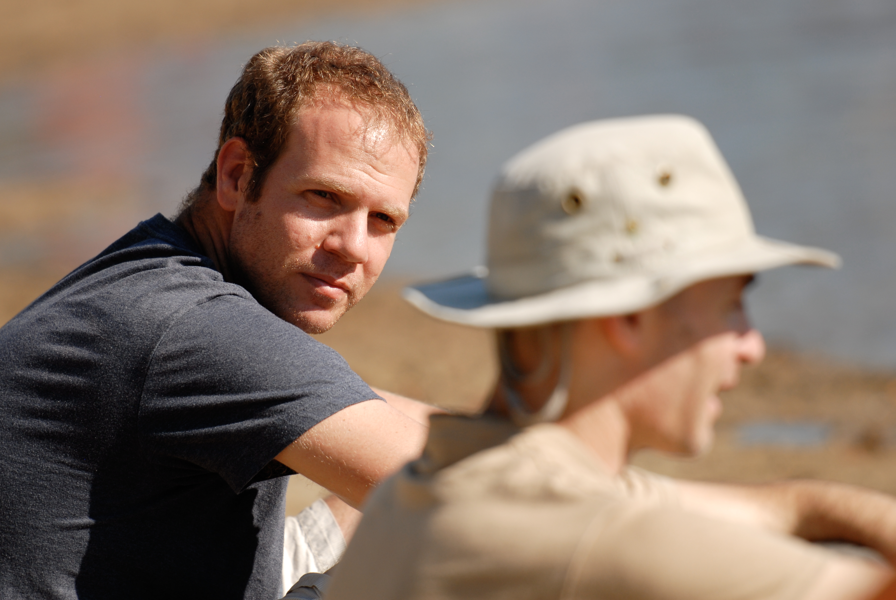 A man in a dark shirt sits outdoors, looking towards the camera, while another person in a hat talks nearby.