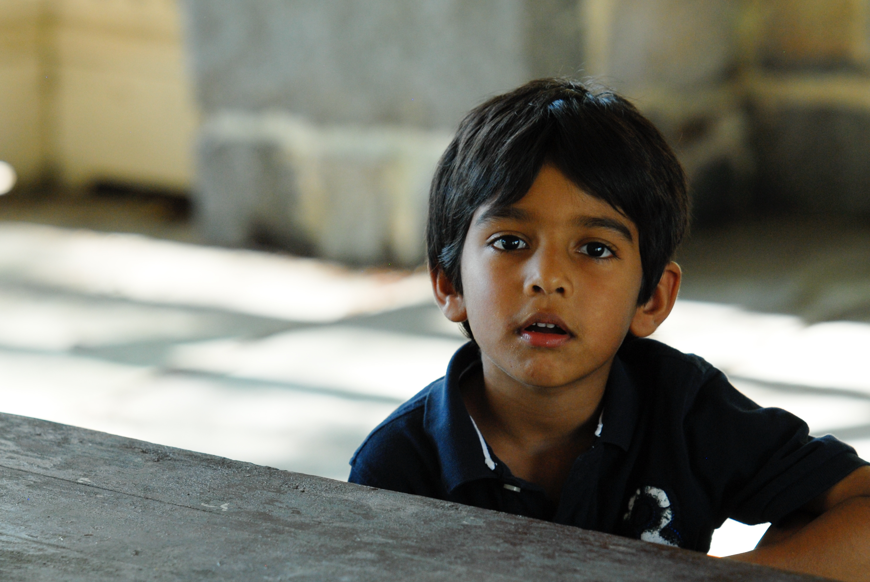 A young boy in a dark shirt sits at a table, looking ahead with a curious expression.