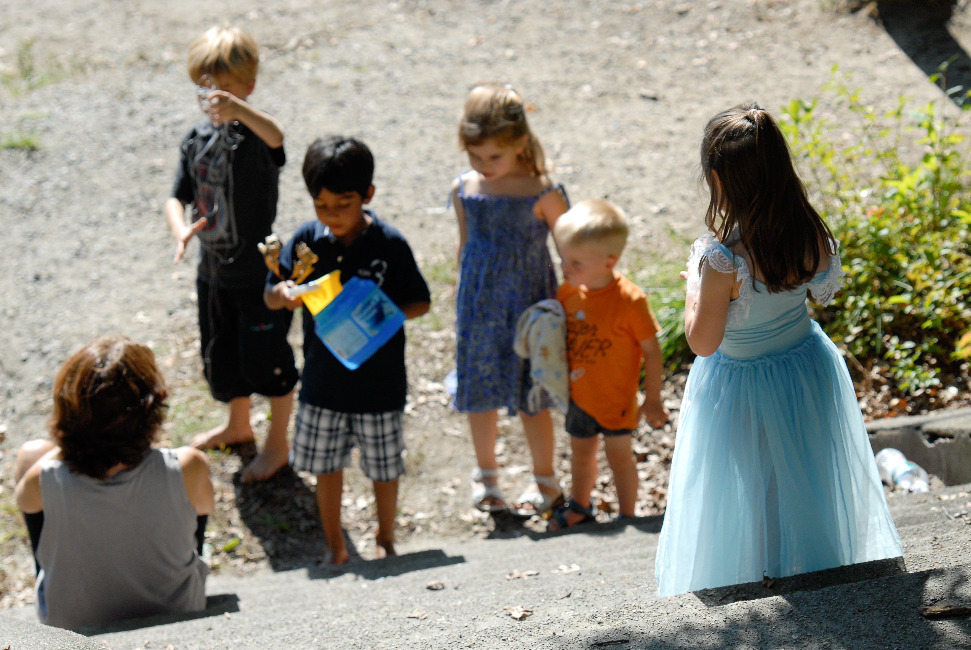 A group of children in colorful outfits stand outside, interacting, while an adult sits on nearby steps.