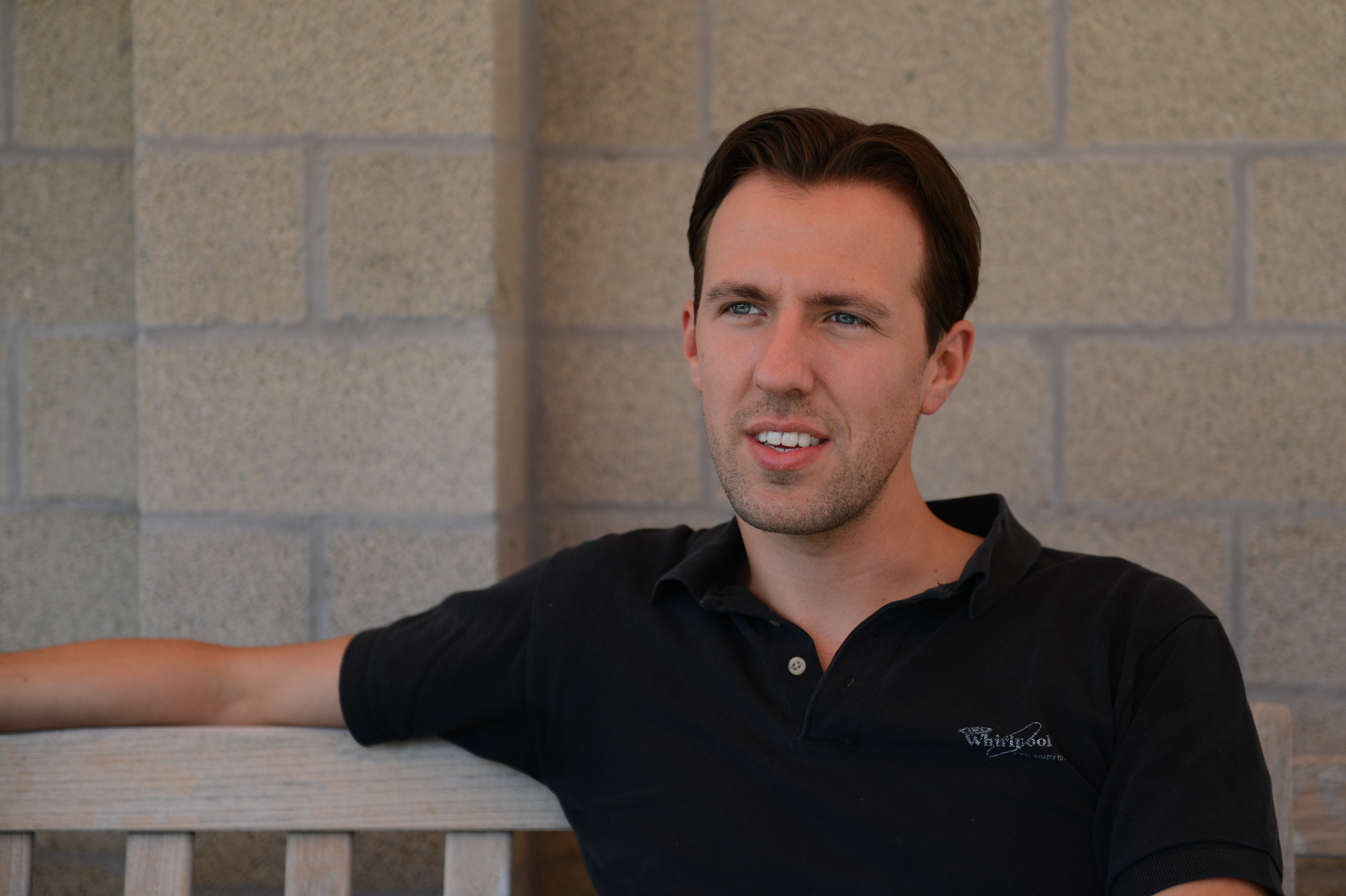 A man in a black polo shirt sits on a bench, leaning back and talking.