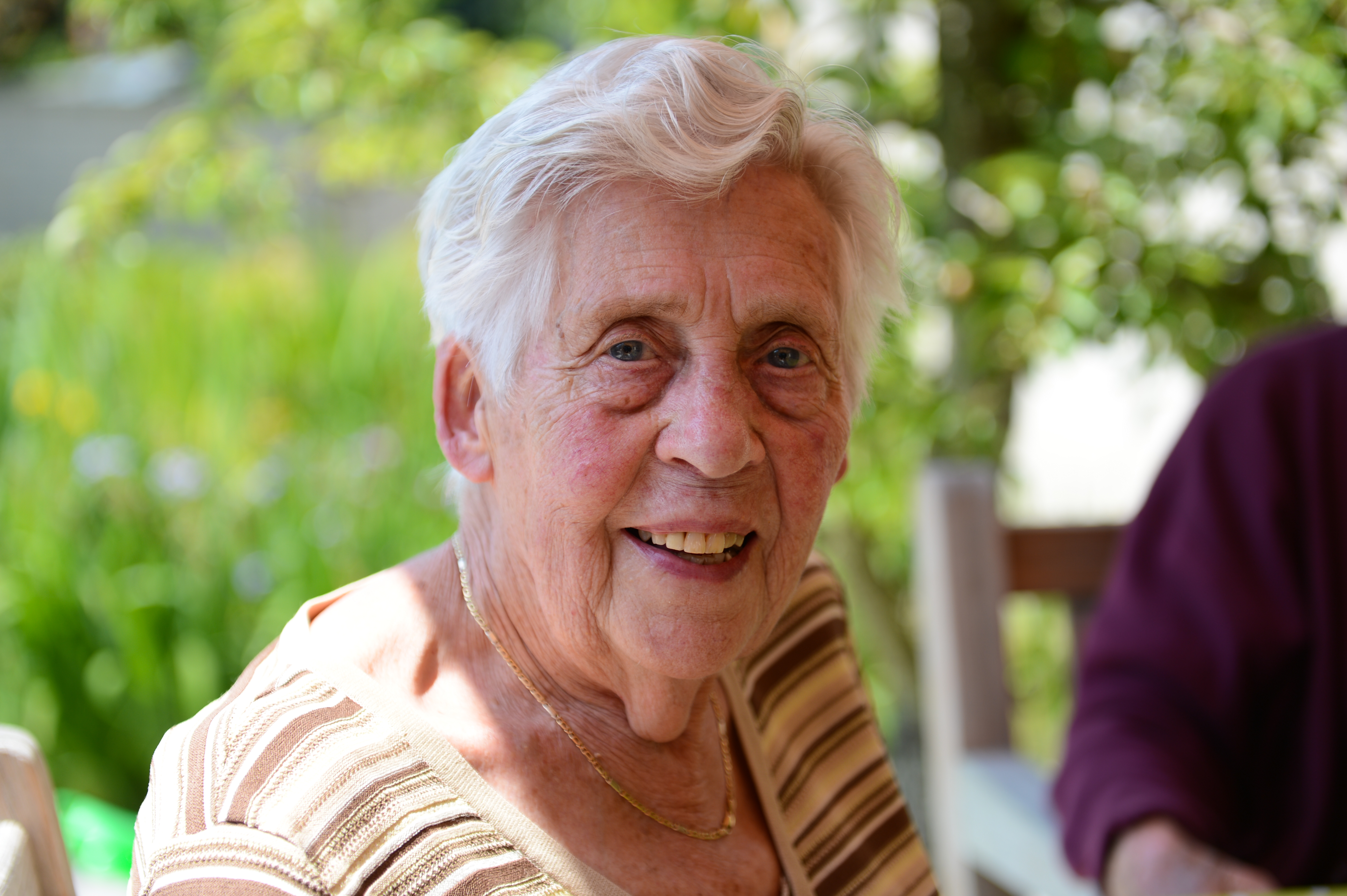 An elderly woman smiles while sitting outdoors at a party, with another person partially visible beside her.