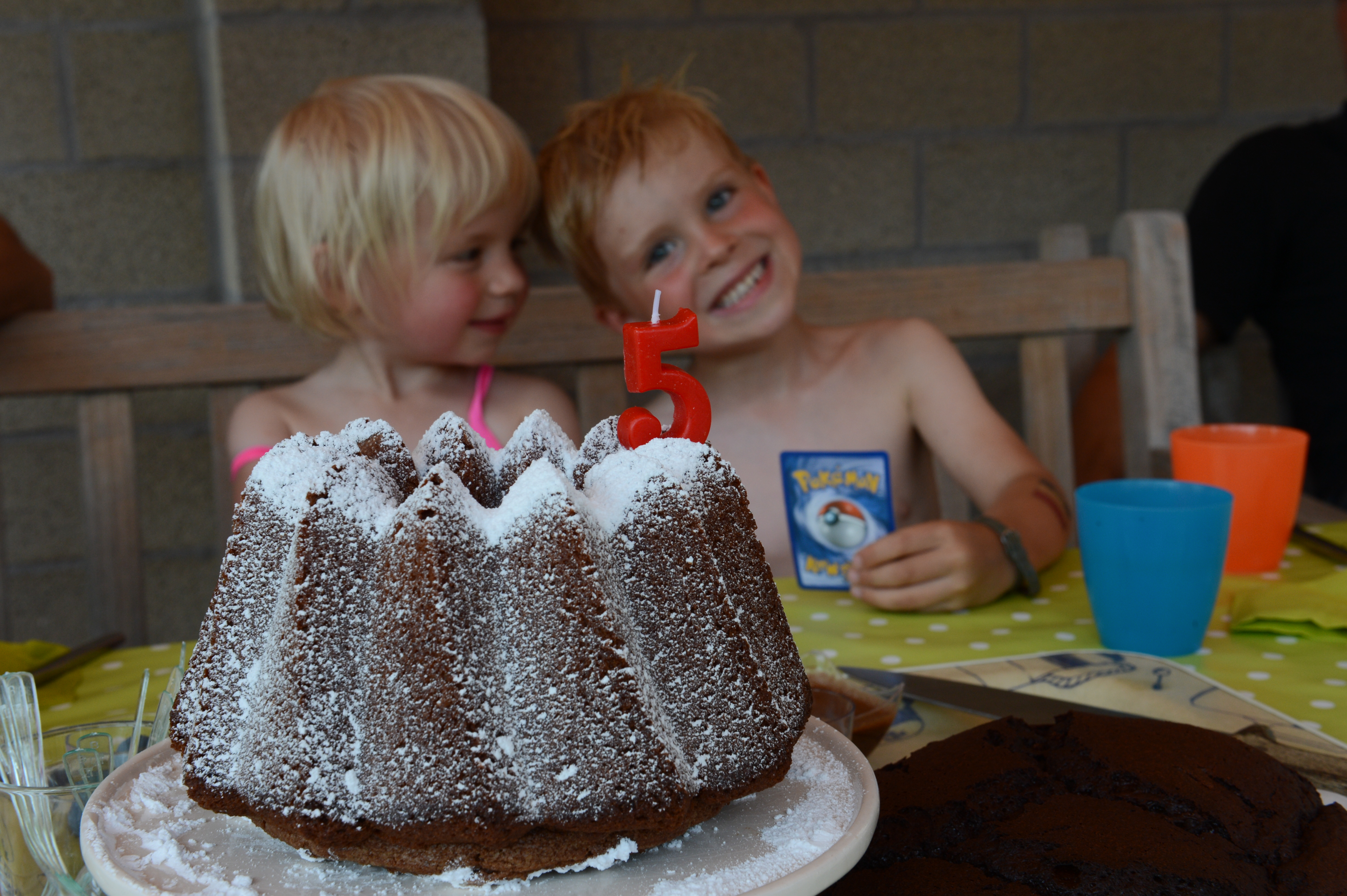 A chocolate cake with a number 5 candle sits on a table, while two children smile in the background.