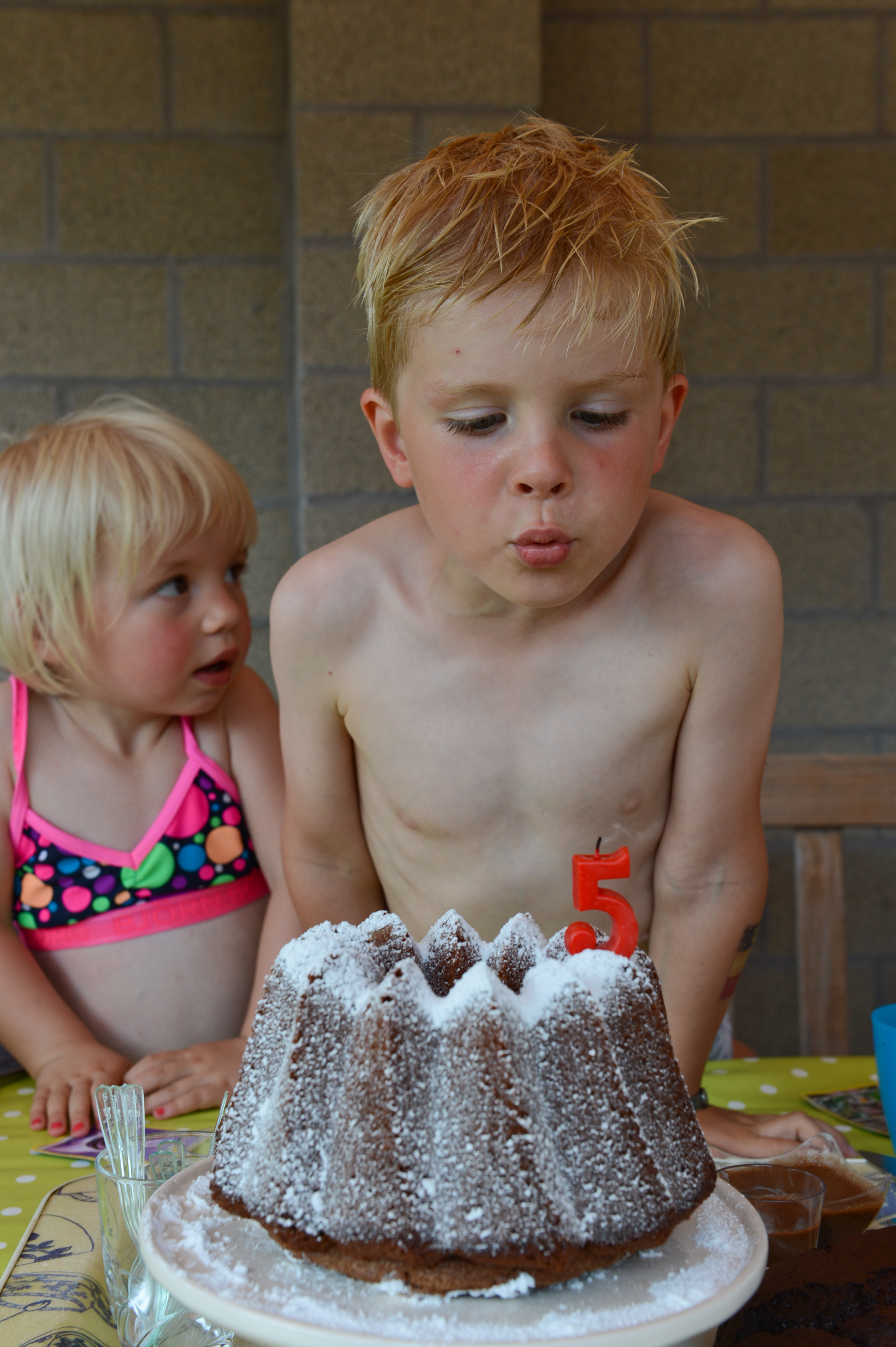 A young boy blows out a number five candle on a cake while a girl watches nearby.