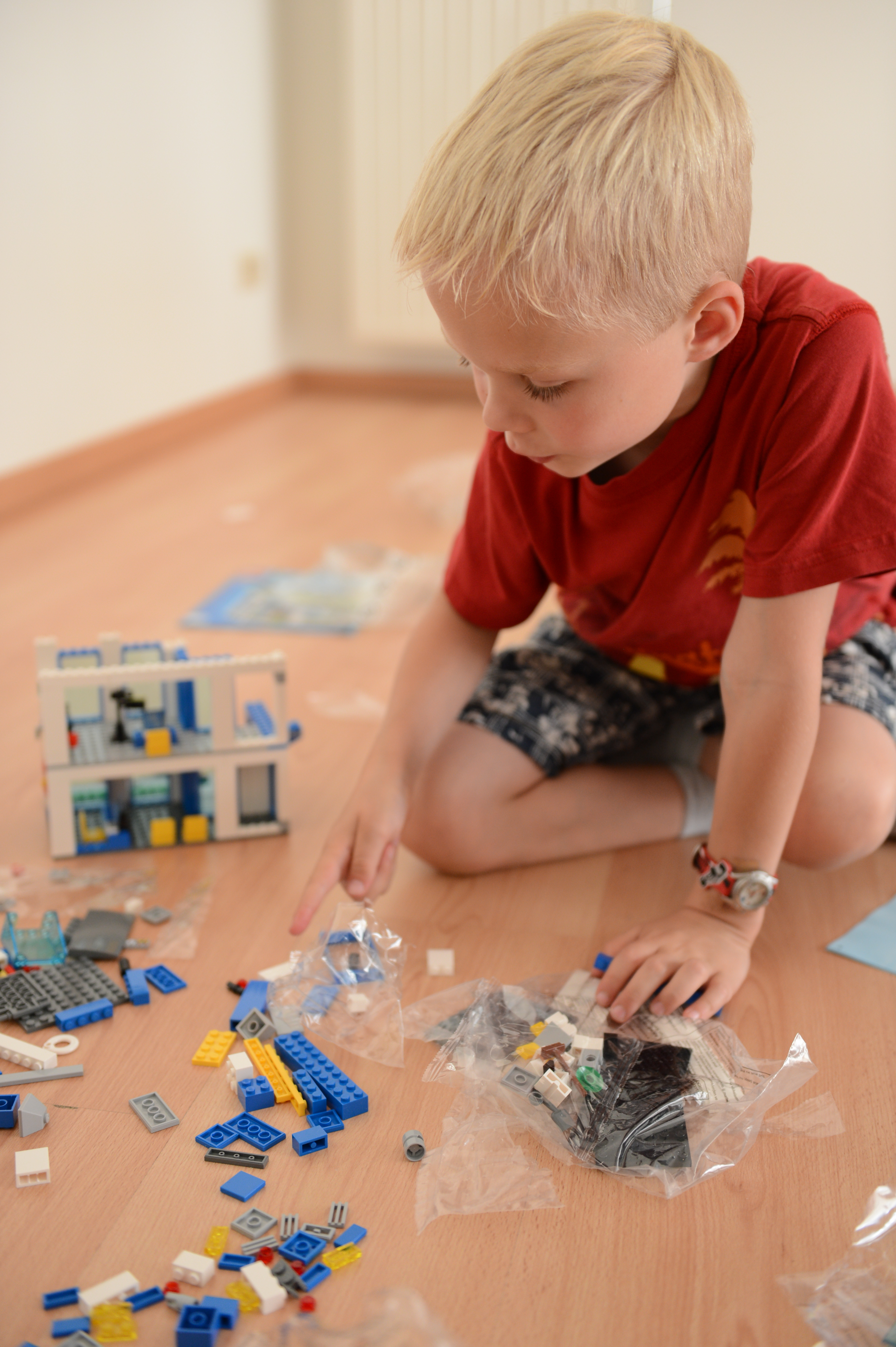A child in a red shirt sits on the floor, sorting and assembling LEGO pieces from plastic bags.