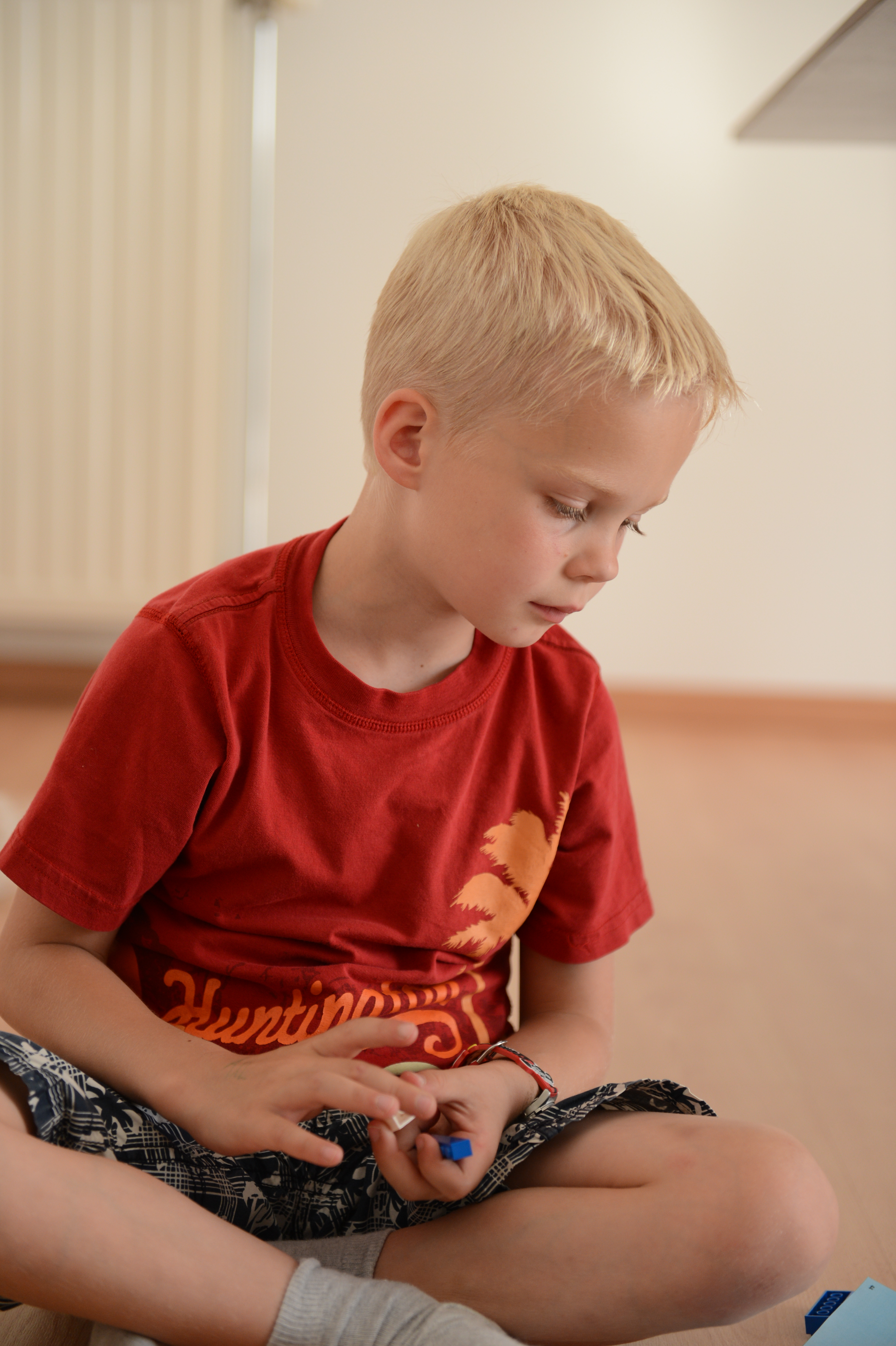 A young child in a red shirt sits on the floor, holding small plastic pieces and looking down.