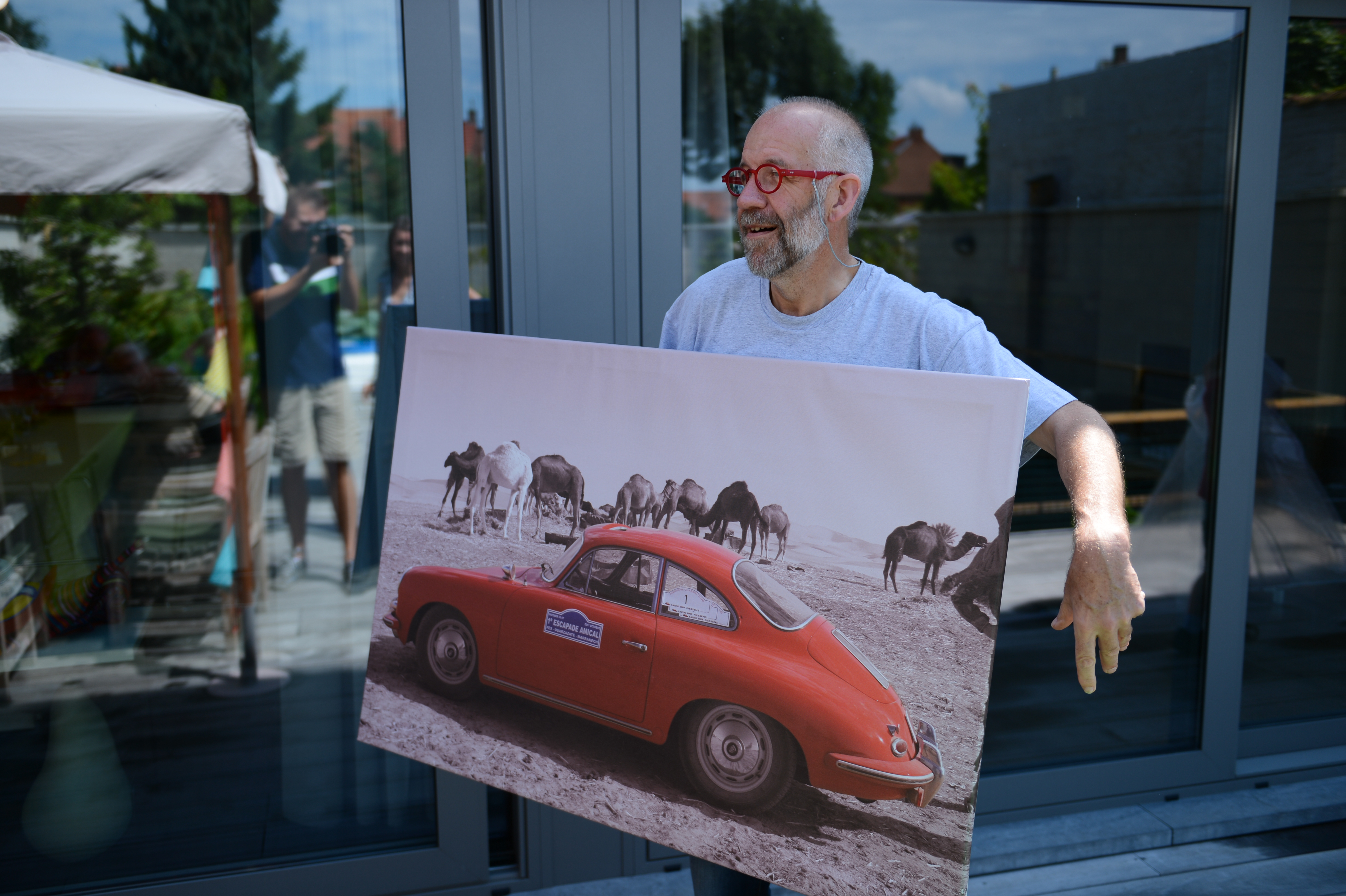 A man with red glasses holds a large framed photo of a red car and camels, smiling outside.