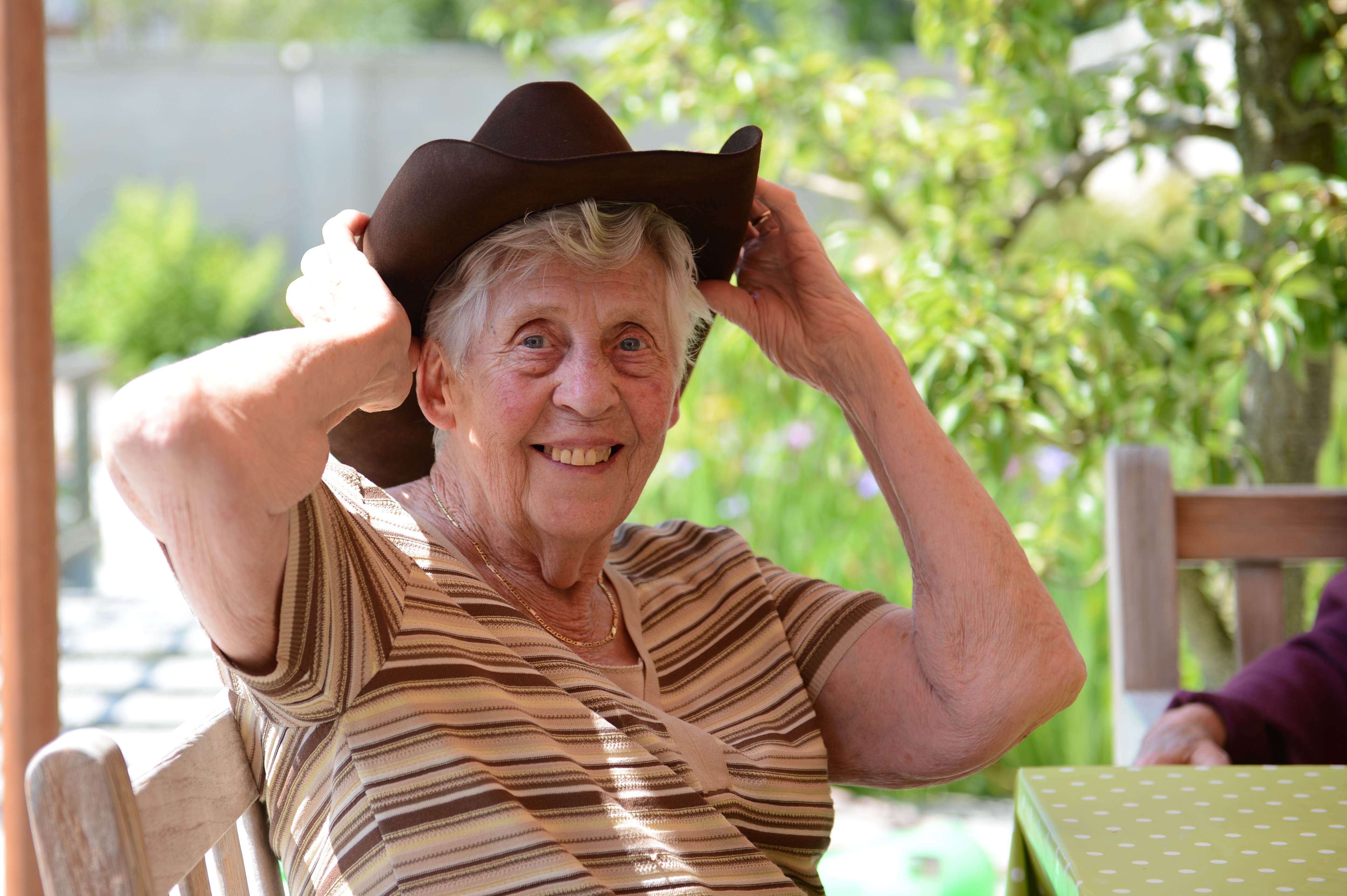 An elderly woman smiles while putting on a cowboy hat at an outdoor party.
