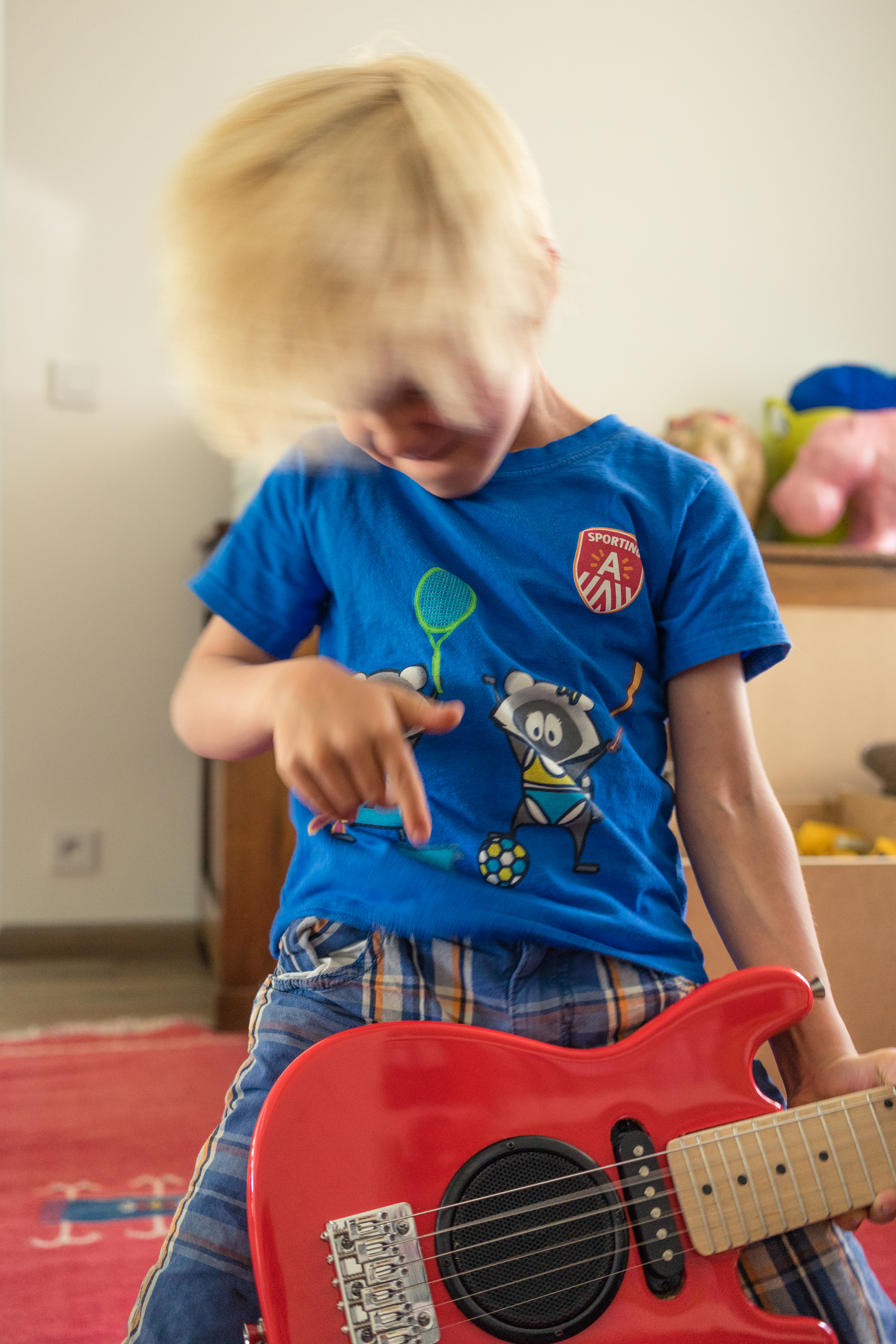 A child in a blue shirt plays a red toy guitar, shaking their head with energy.