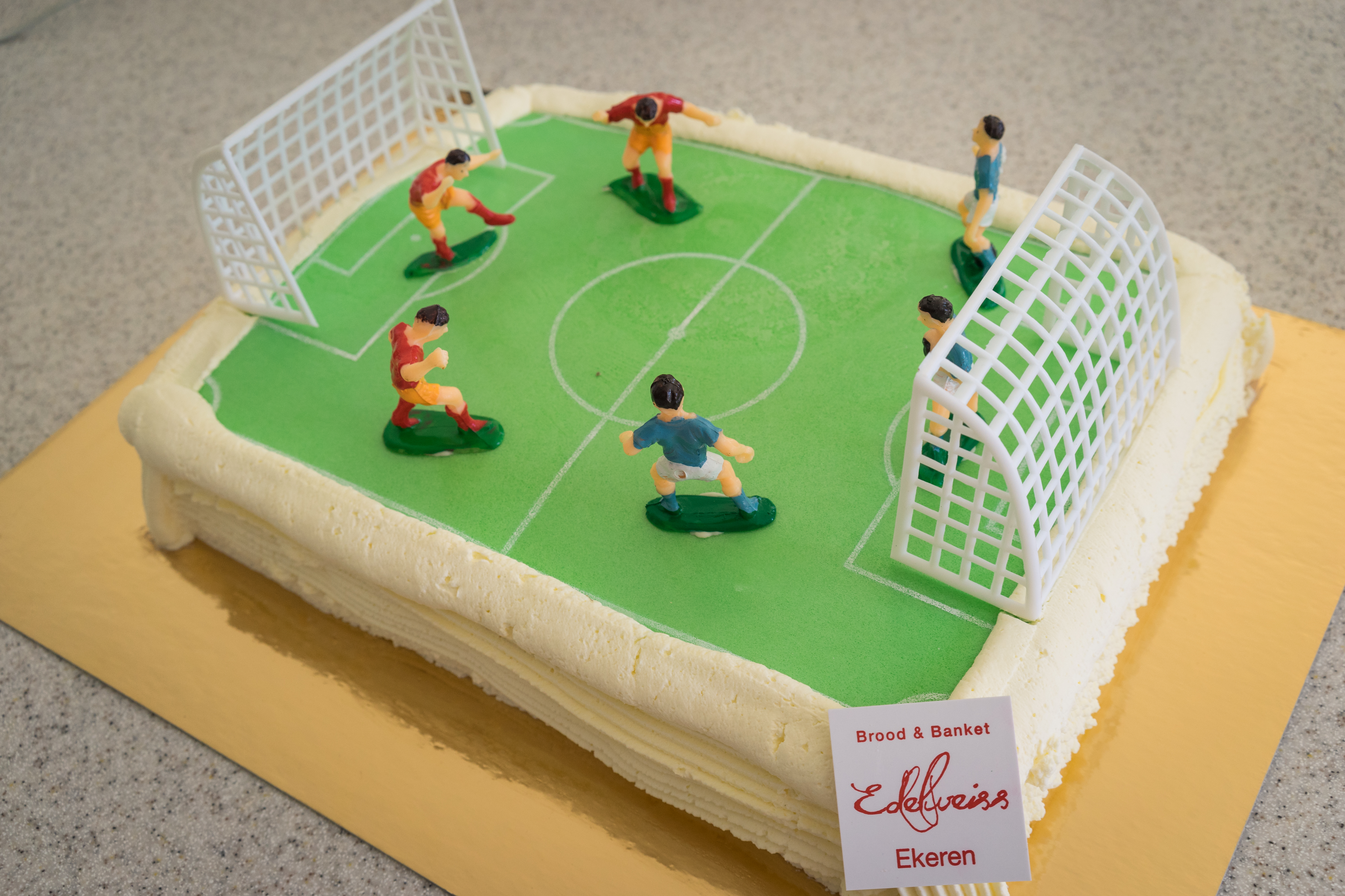 A rectangular cake decorated as a soccer field with small player figurines and goals on top.