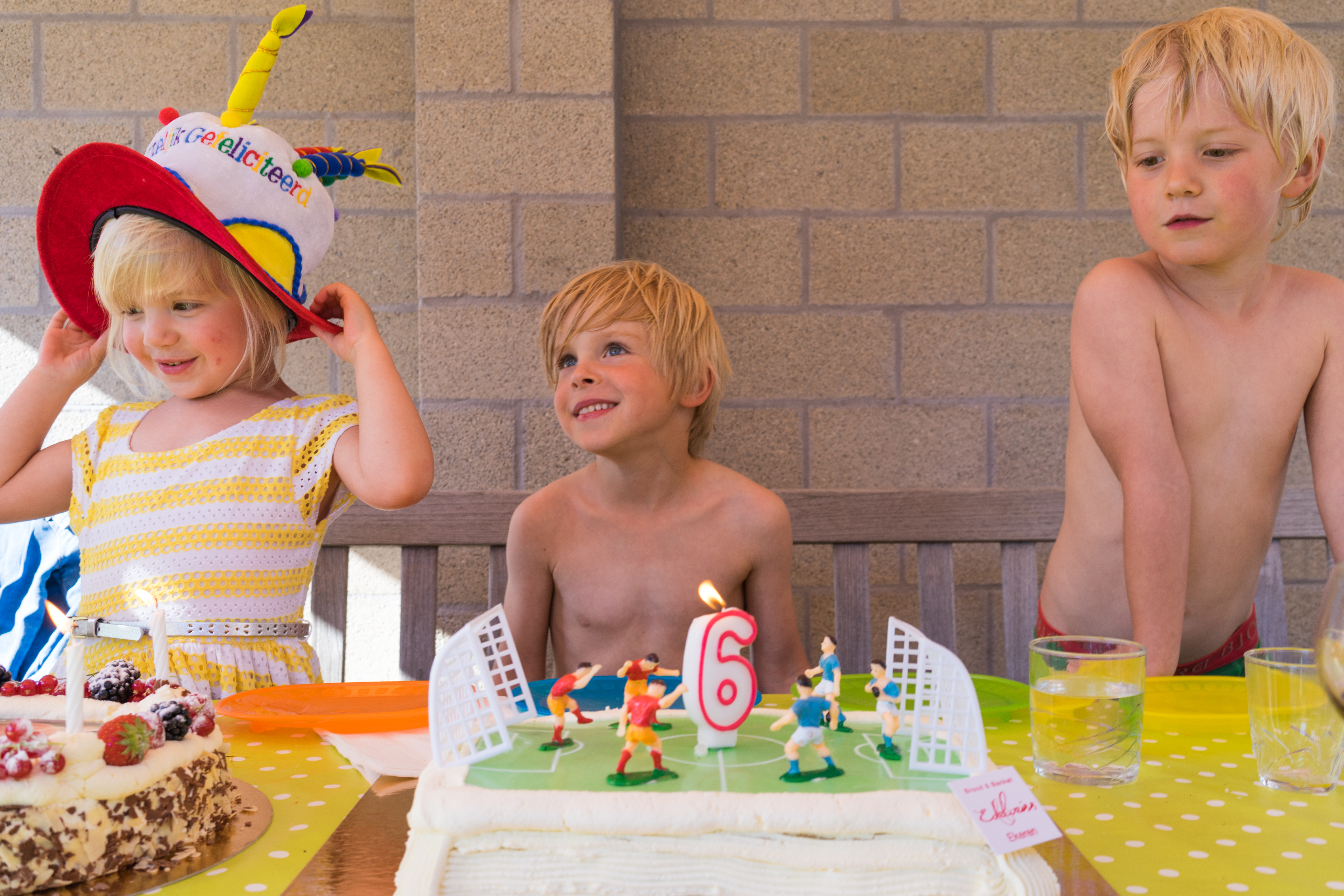 Three children gather around birthday cakes, one with a number six candle, while a girl adjusts a festive hat.