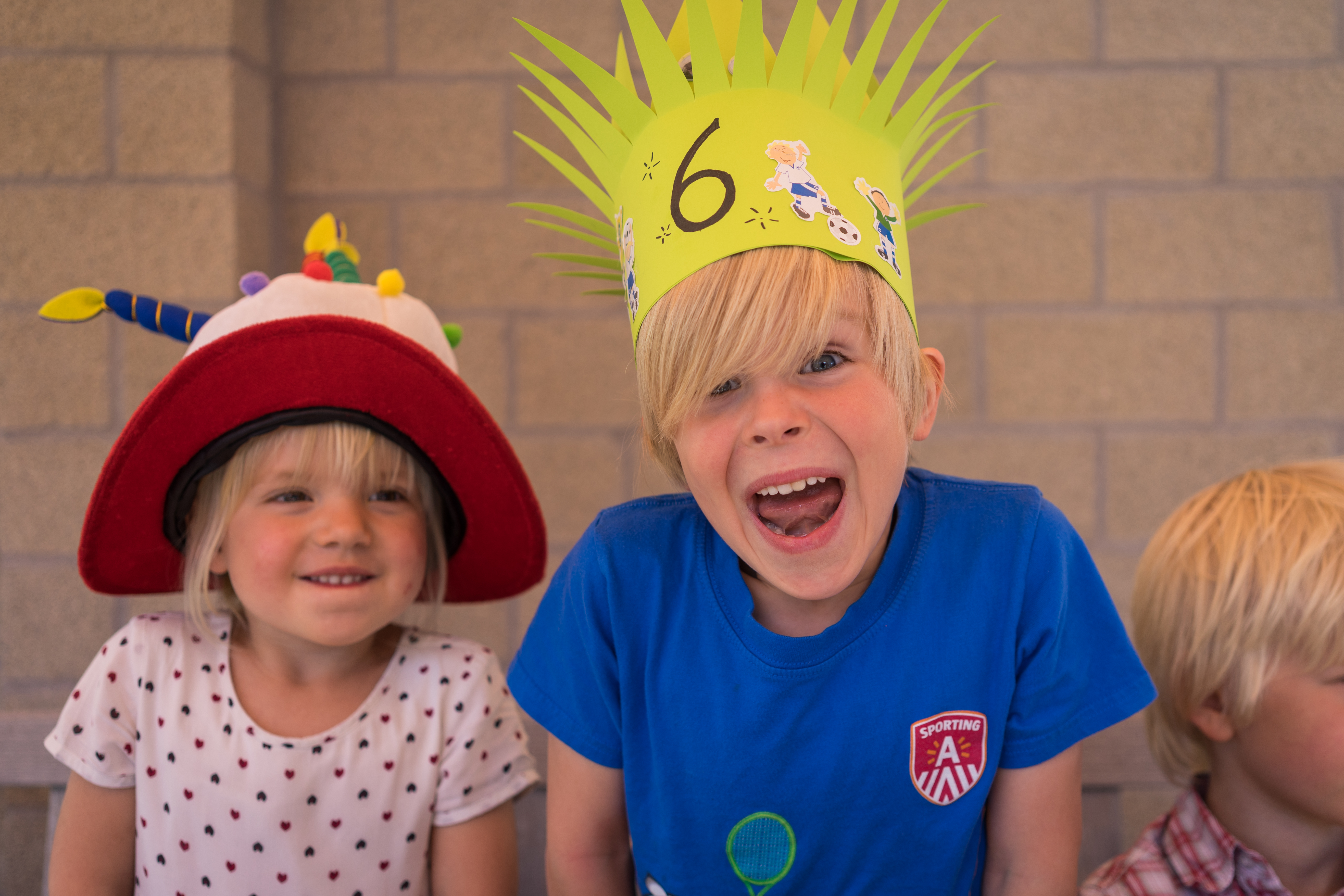 Two smiling children wear festive hats, with one excitedly showing a handmade crown labeled "6" at a birthday party.