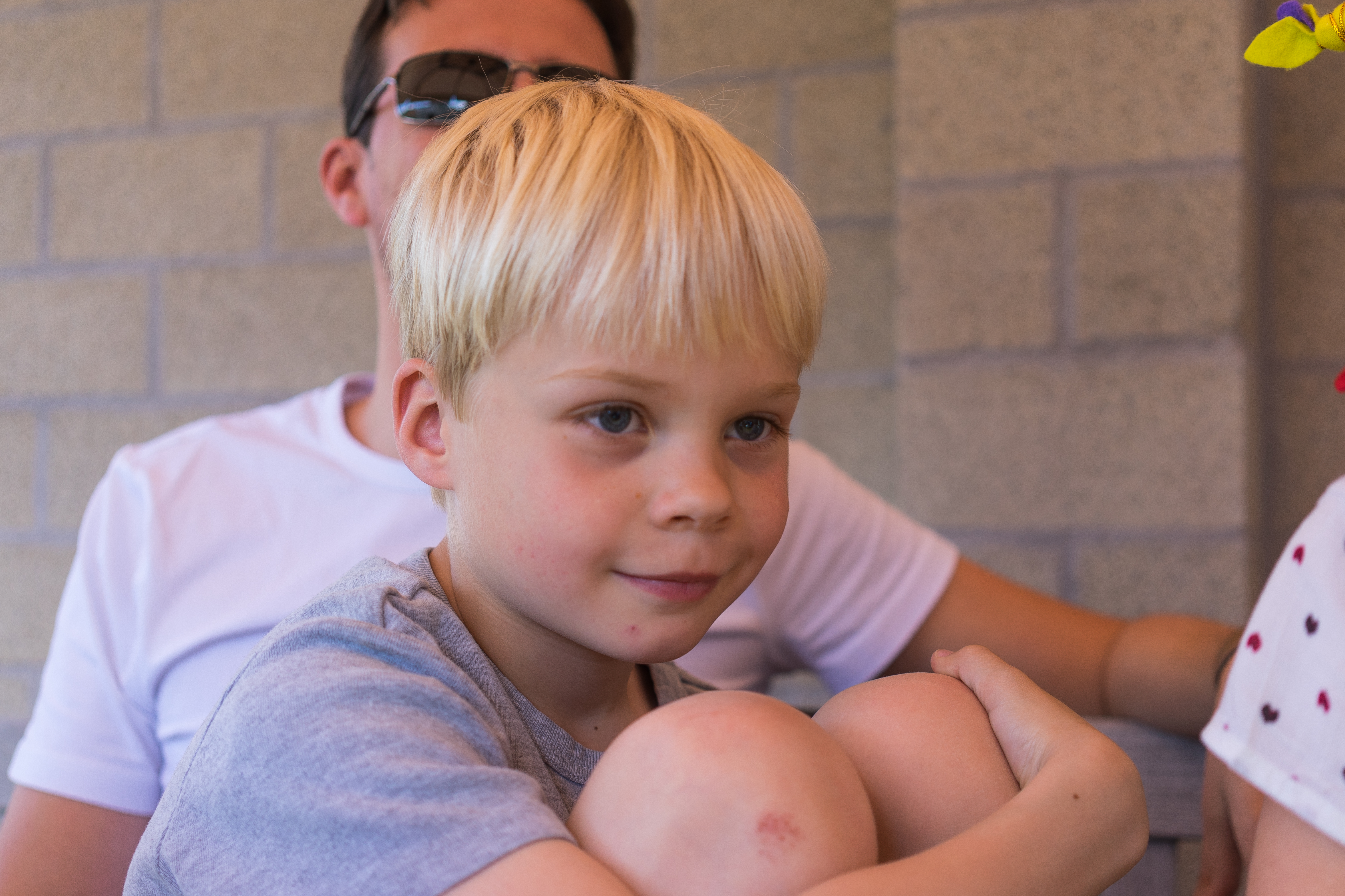 A young boy with blonde hair sits with his knees up, looking ahead, while an adult sits behind him.