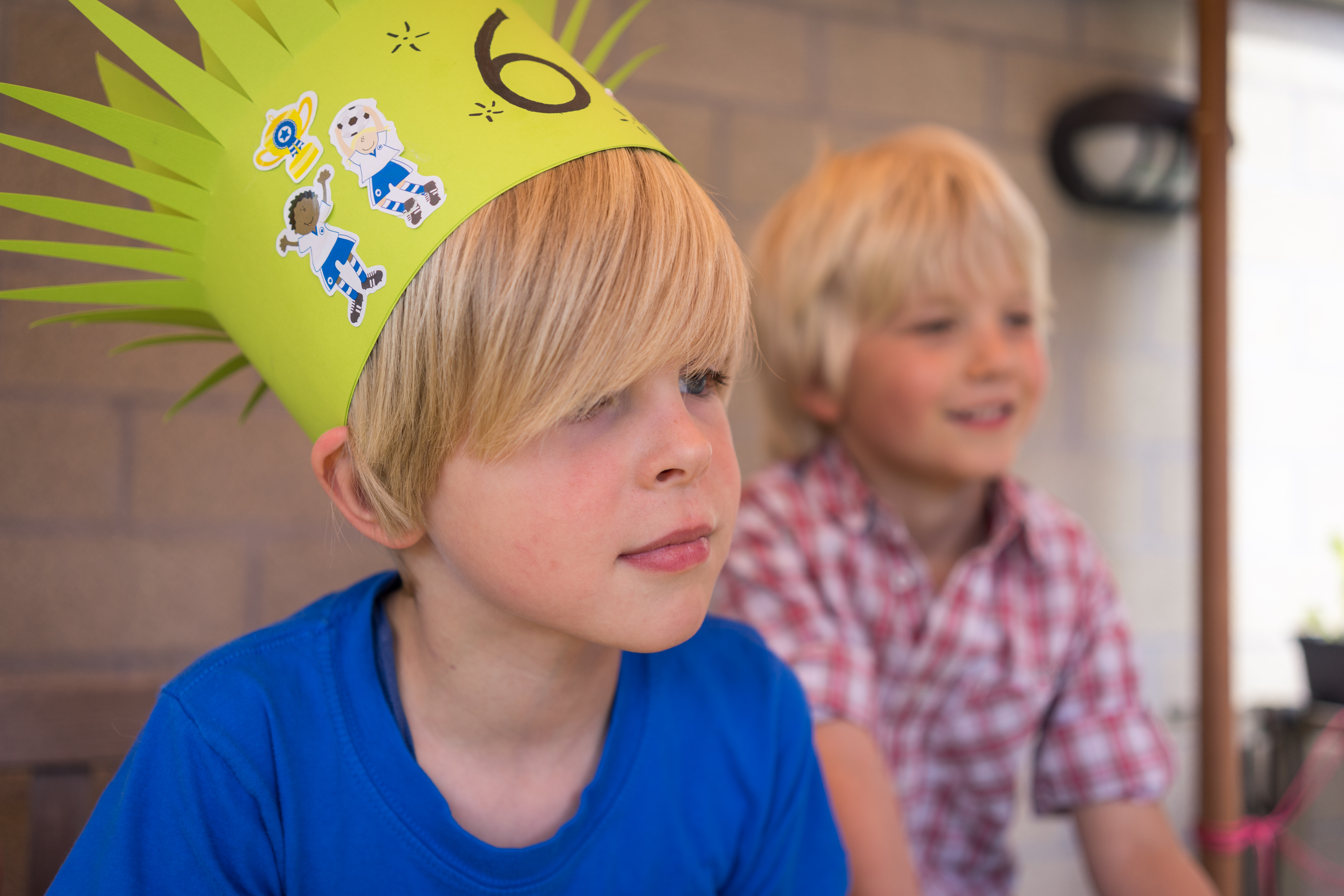A child wearing a handmade birthday crown sits attentively, while another child in a plaid shirt smiles behind him.