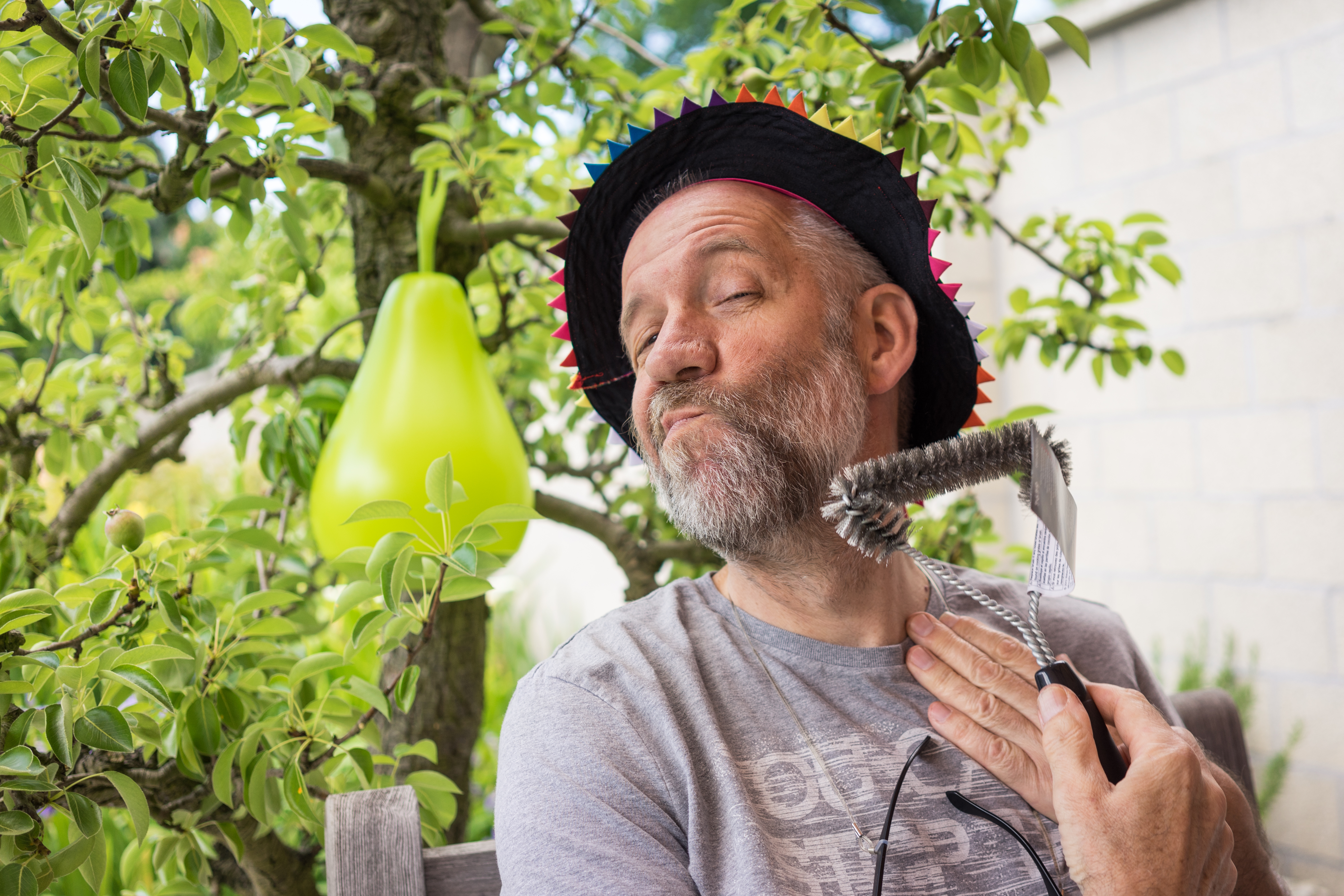 A man wearing a festive hat playfully brushes his beard with a grill brush while making a funny expression.
