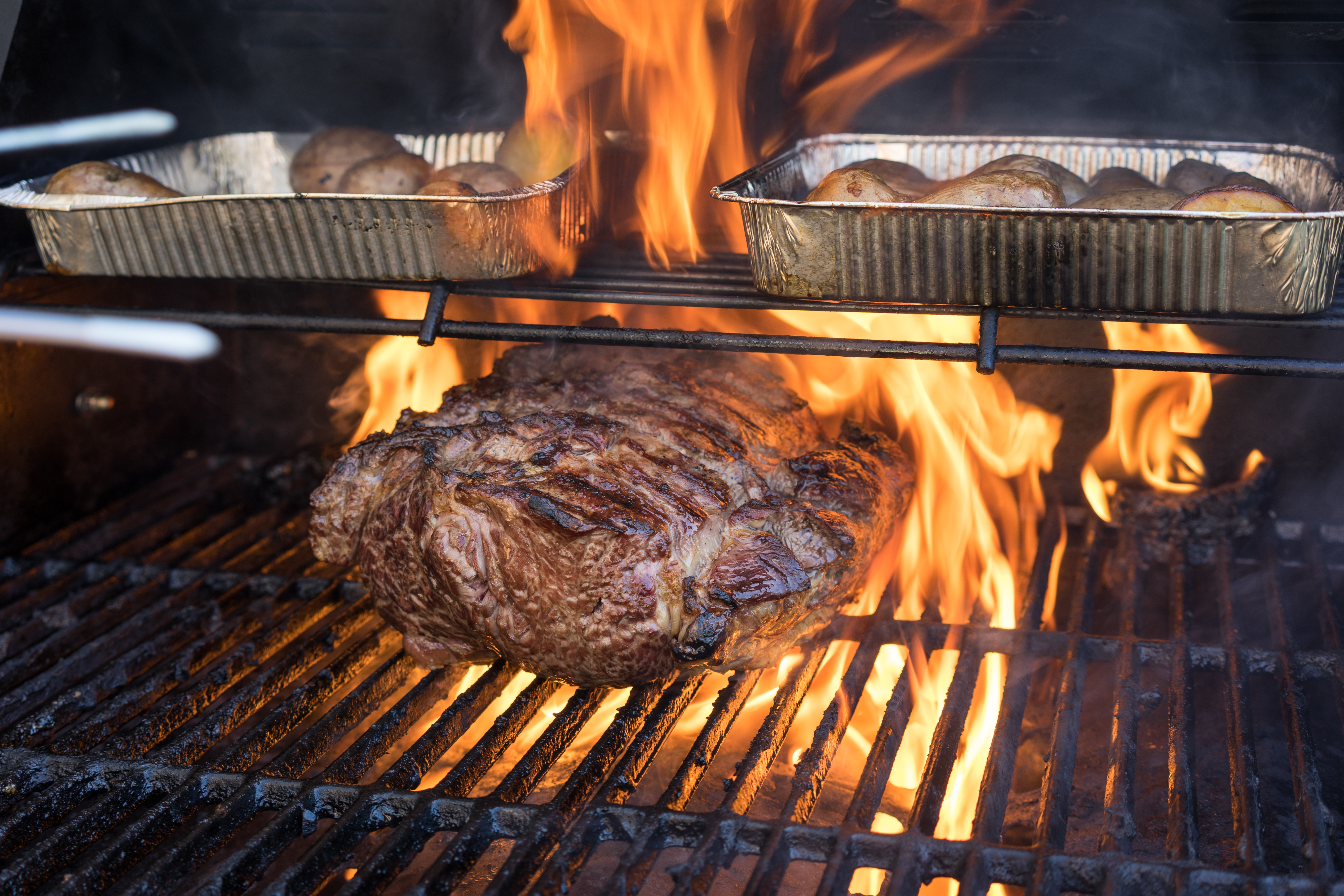 A large piece of meat and trays of potatoes cook on a grill with flames rising.