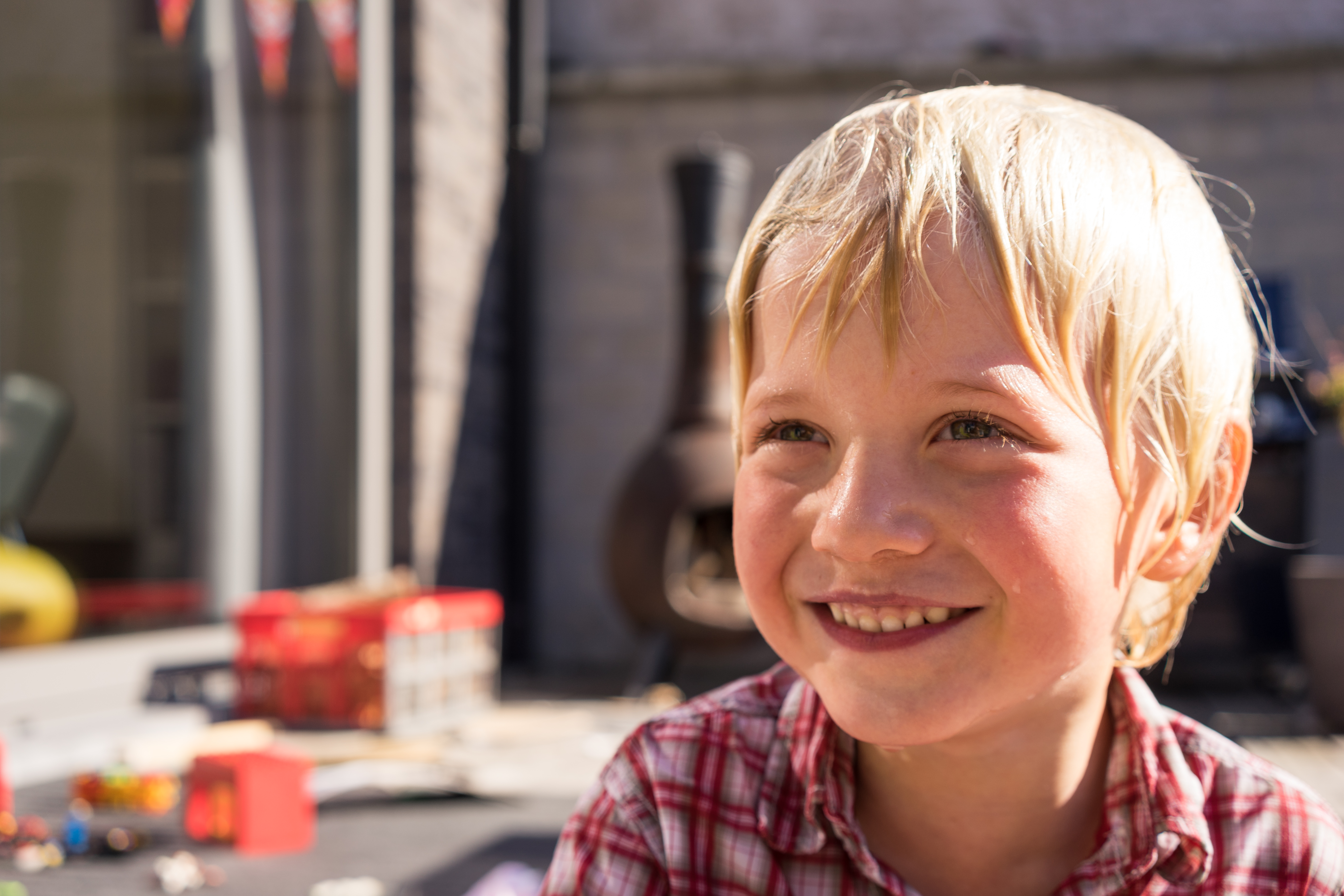 A smiling child in a red plaid shirt enjoys an outdoor birthday party with toys and decorations in the background.