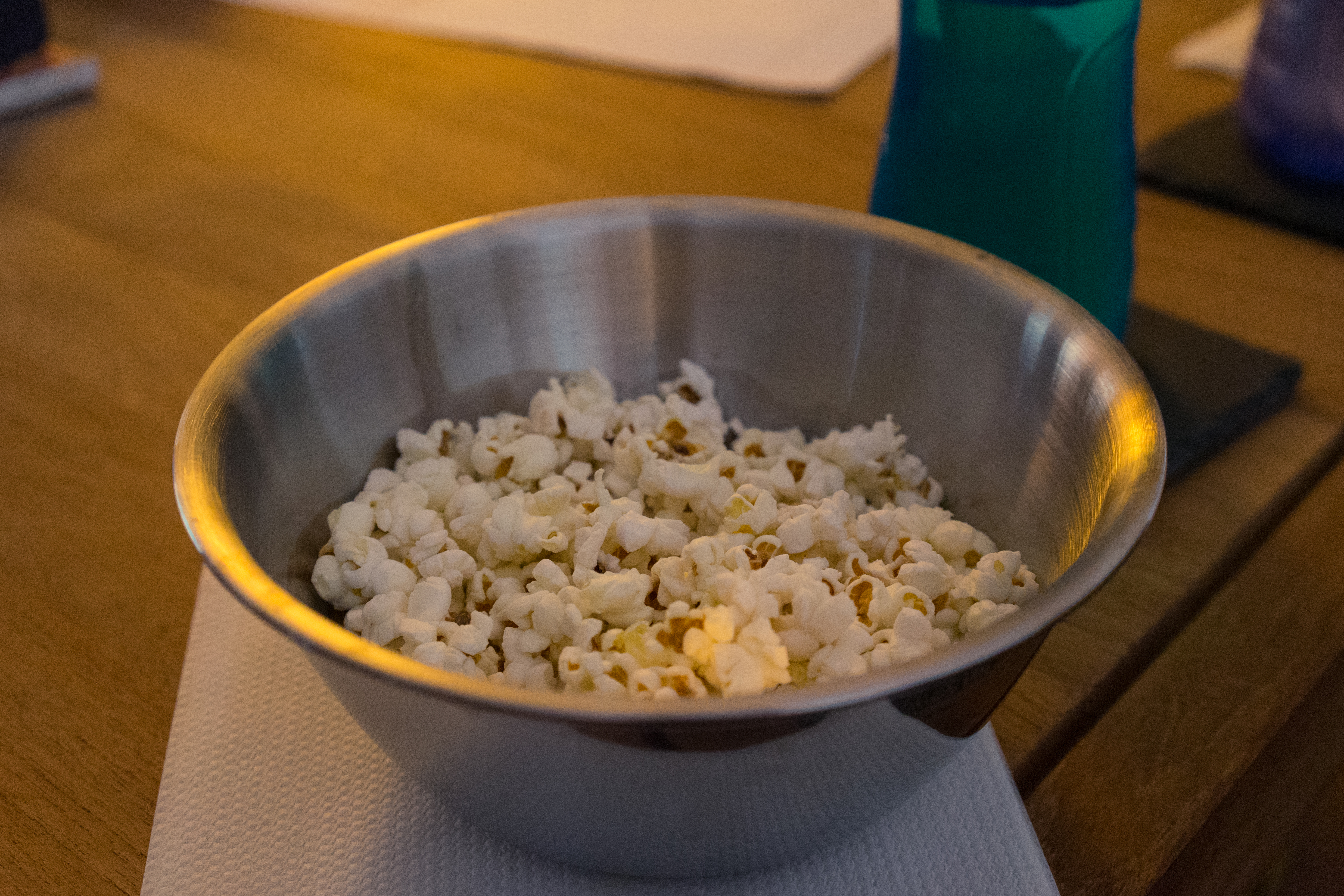 A metal bowl filled with popcorn sits on a wooden table, with drinks nearby.