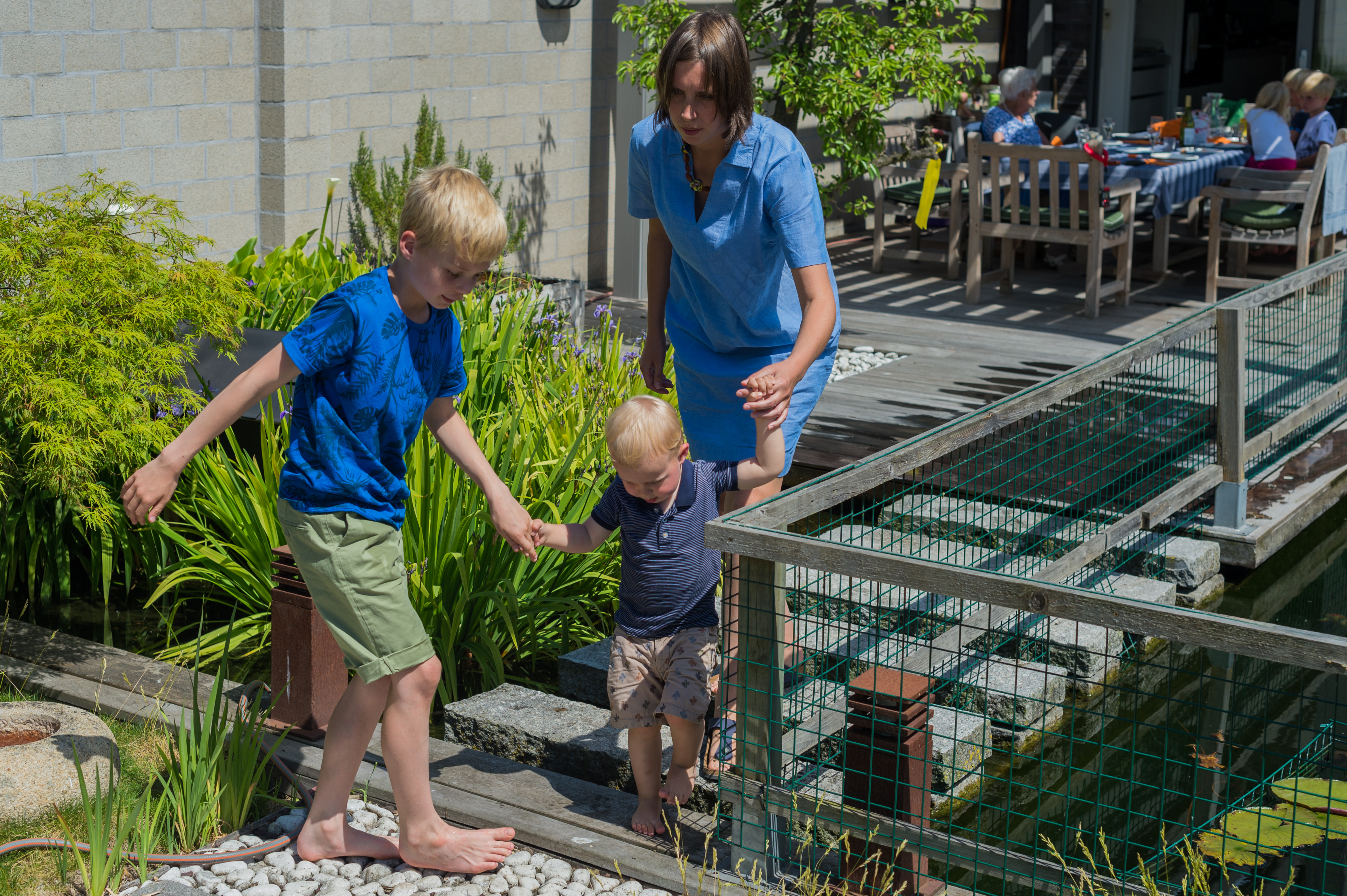 A woman helps two young children walk barefoot on a narrow path beside a small pond in a garden.