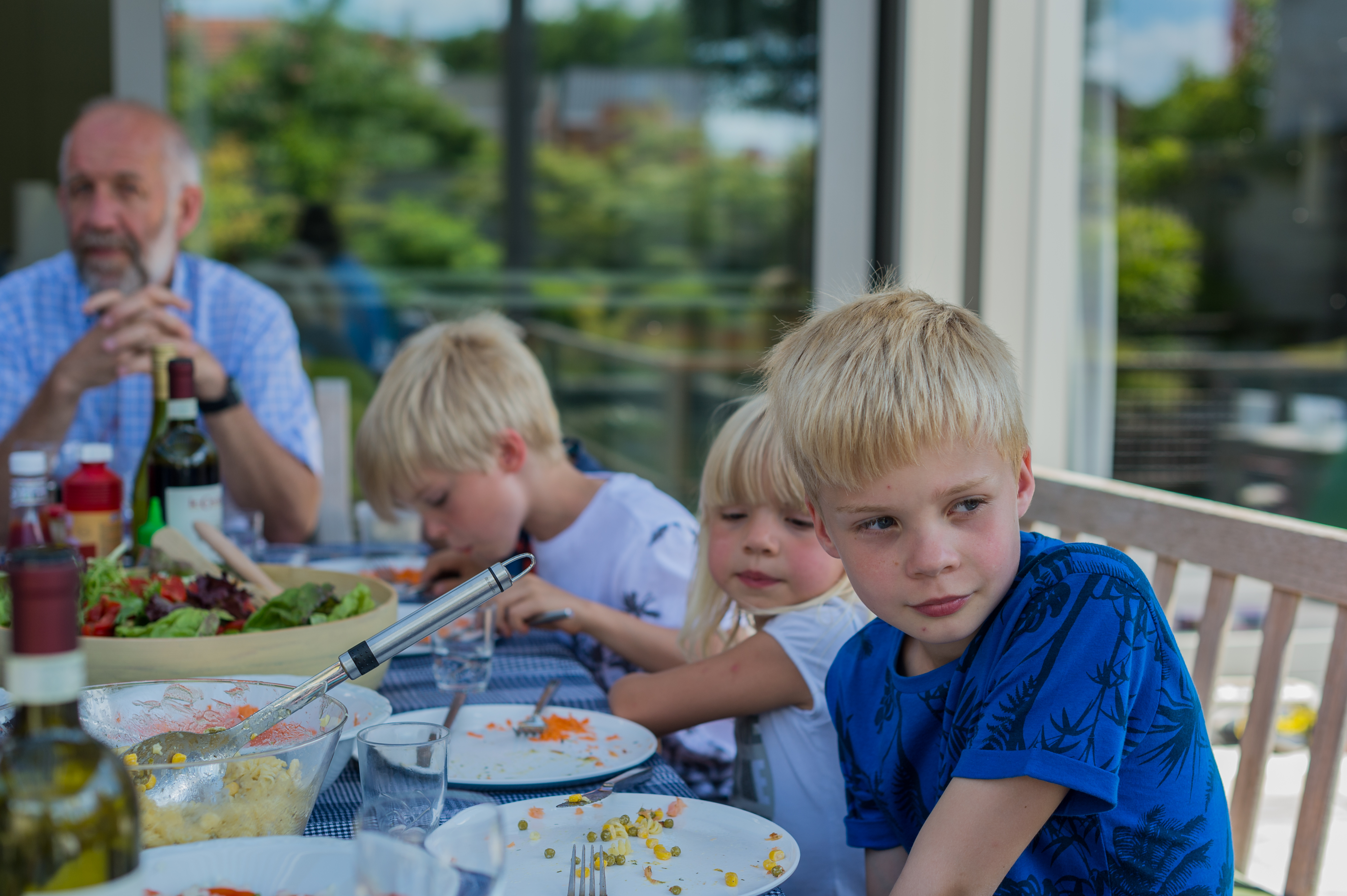 Children and an older man sit at an outdoor table with food, drinks, and salad during a barbecue gathering.