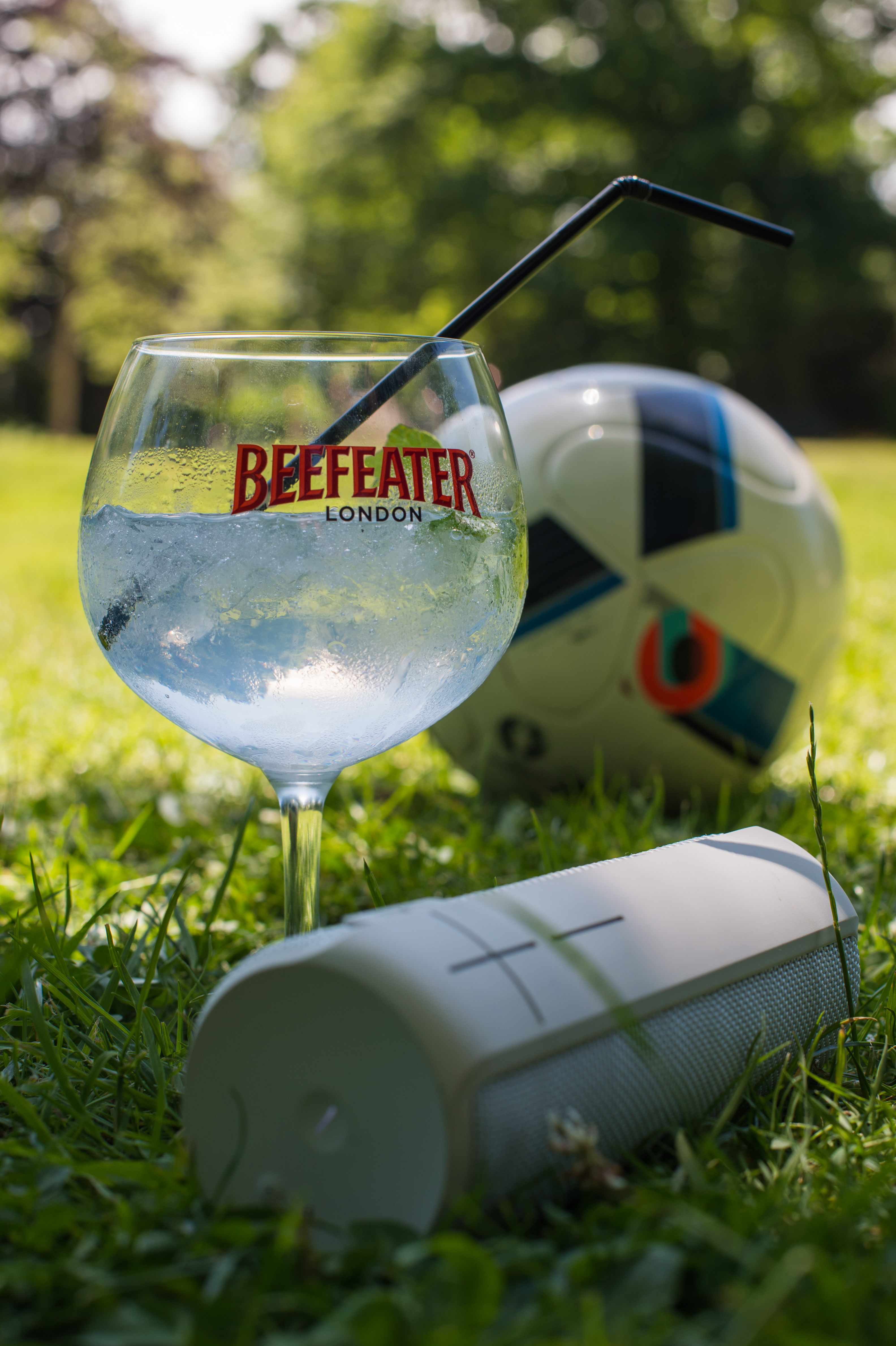 A glass with a drink, a speaker, and a soccer ball rest on the grass in a park.