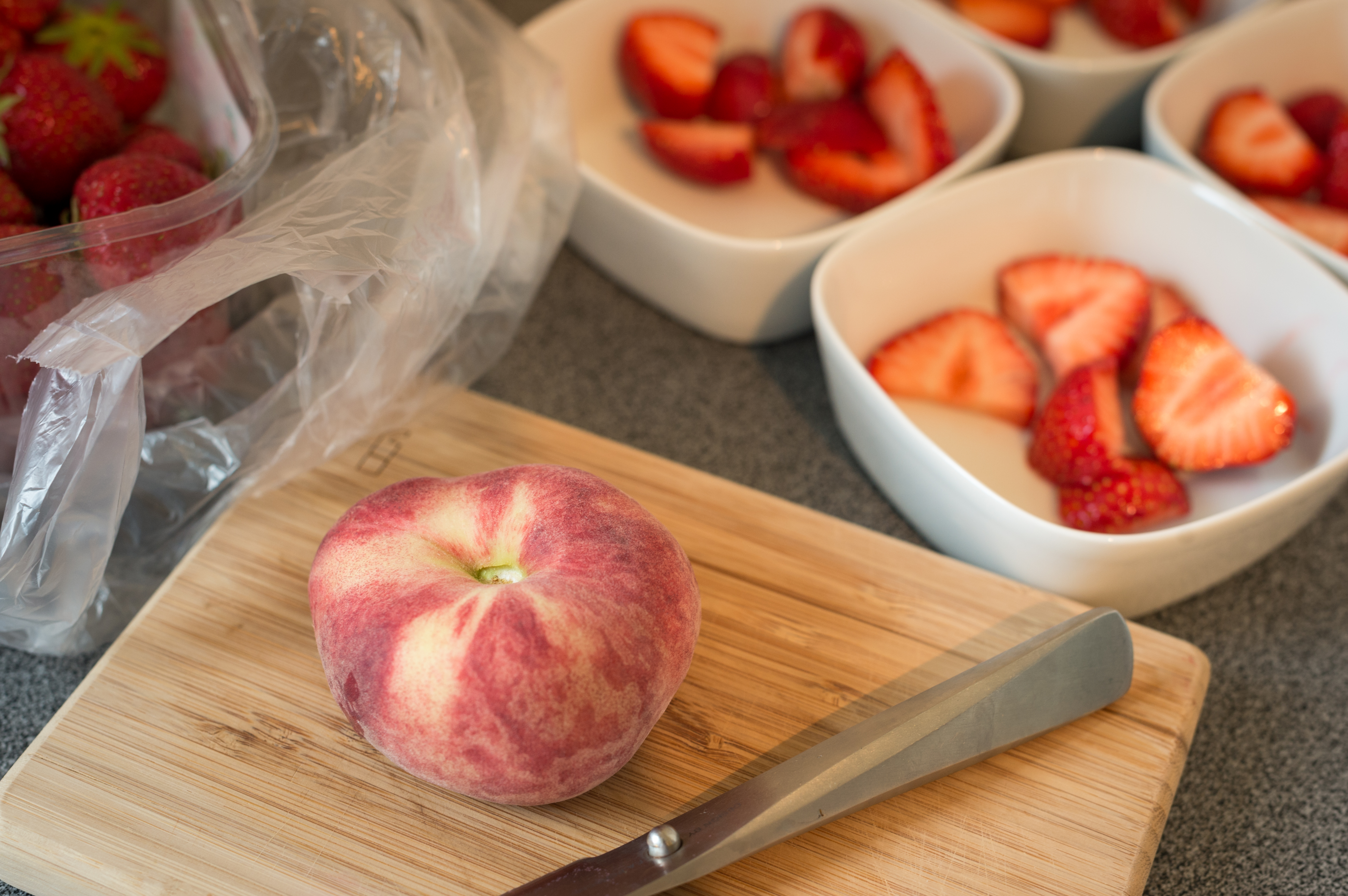 A cutting board with a peach and knife, with bowls of sliced strawberries in the background.