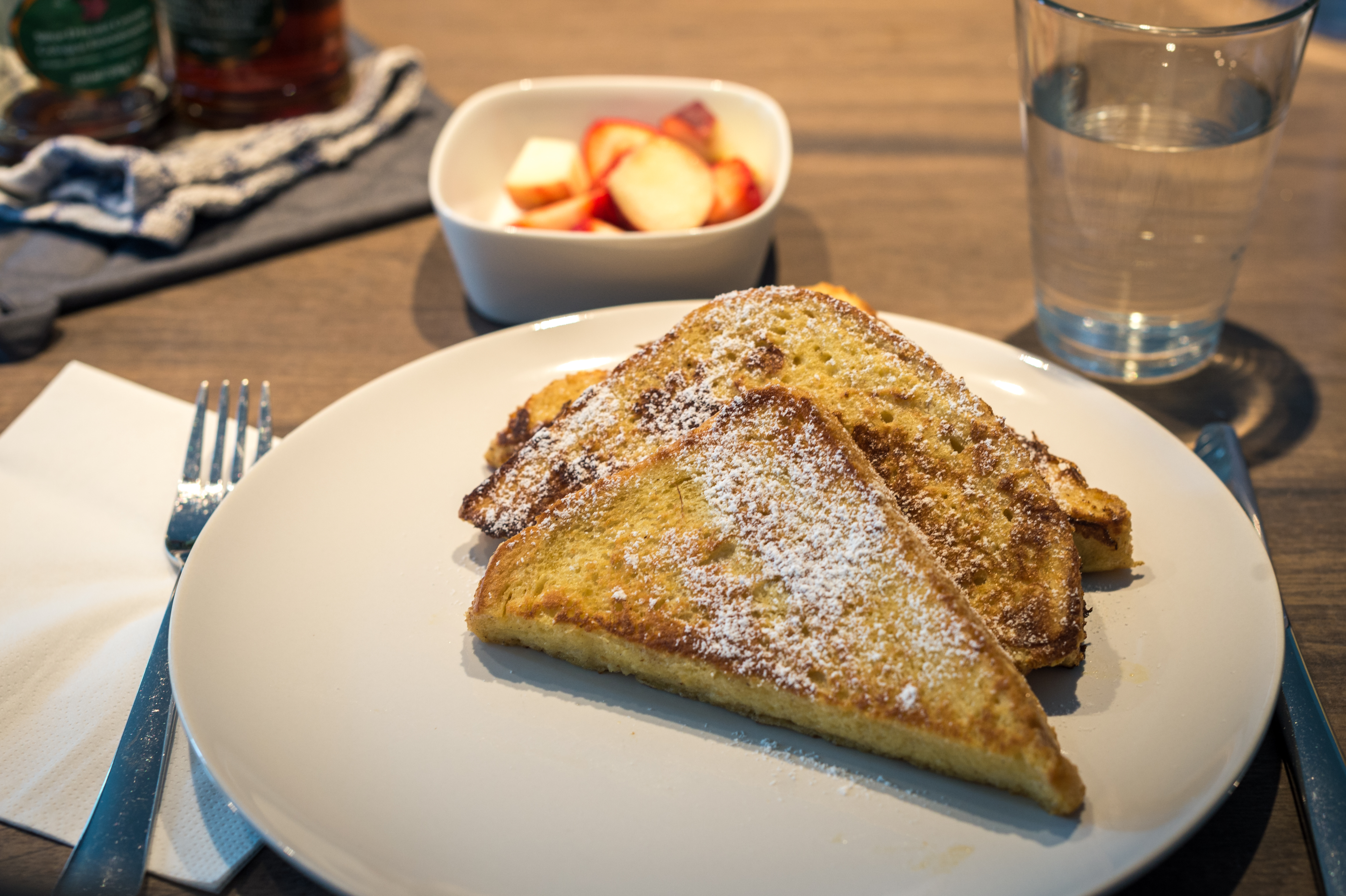 A plate of French toast with powdered sugar, served with a side of fruit and a glass of water.