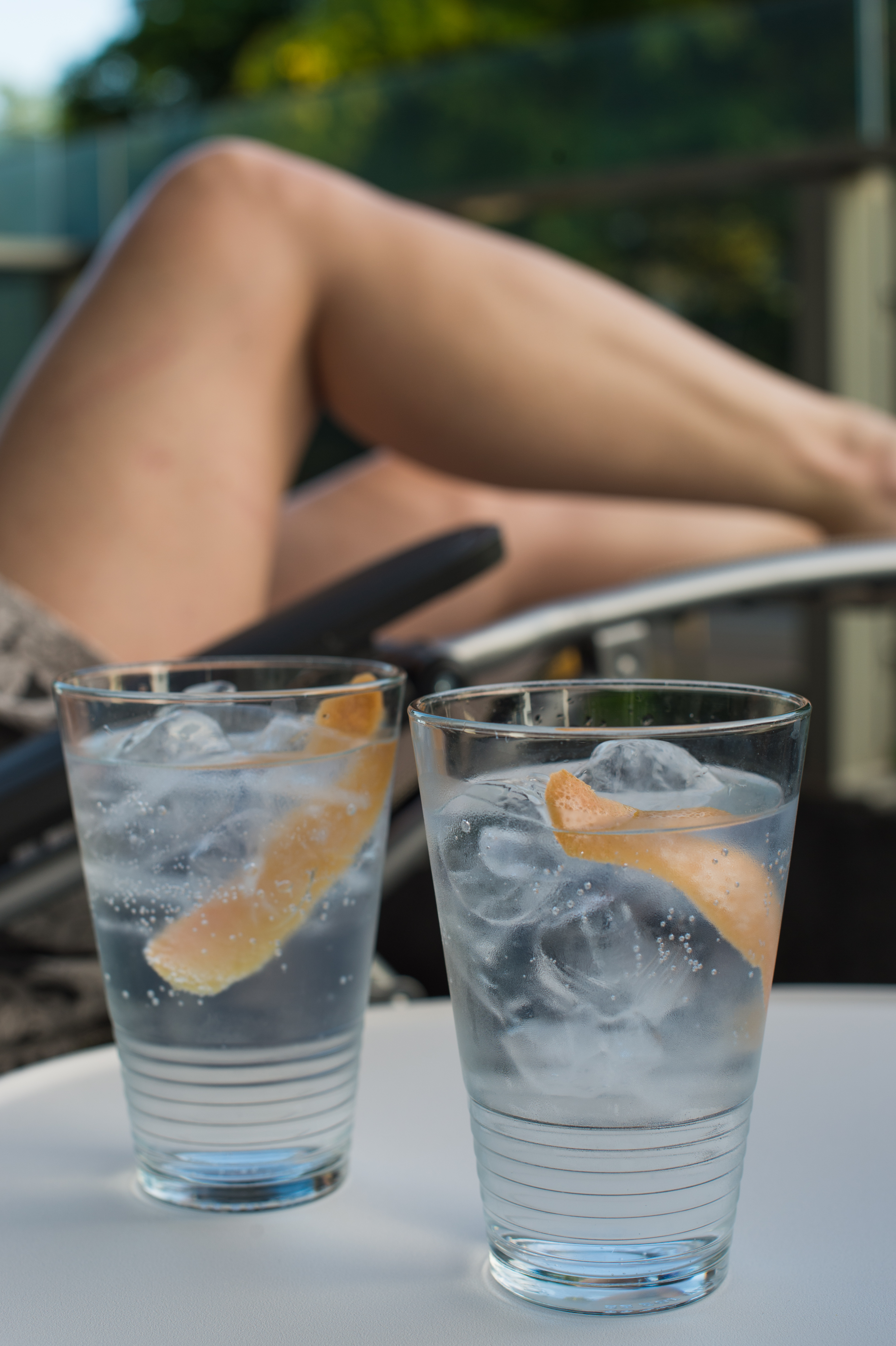 Two glasses of gin and tonic with ice and citrus peel on a table, with legs resting nearby.