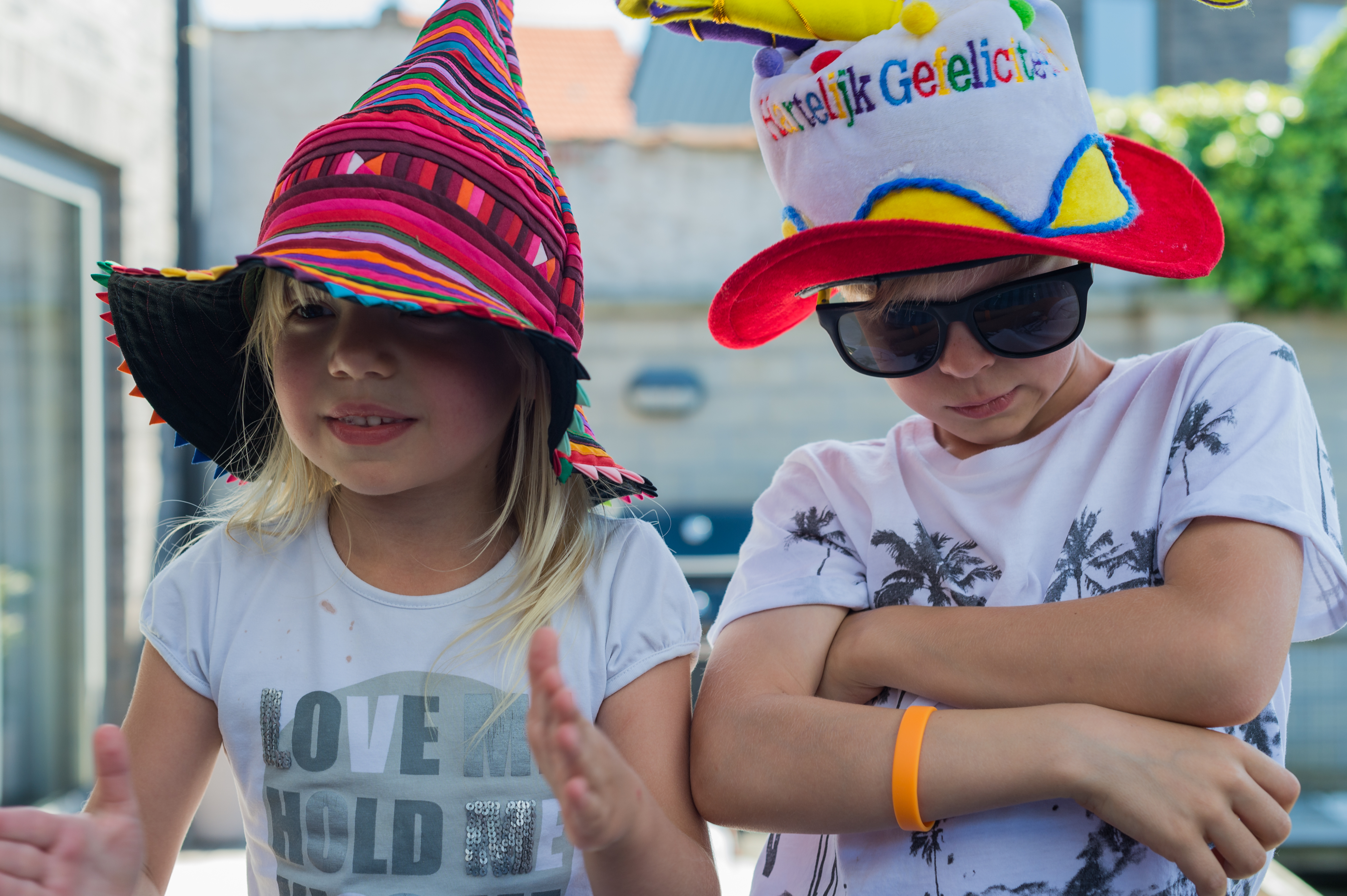 Two children wearing colorful hats pose playfully, one clapping and the other crossing their arms with sunglasses on.