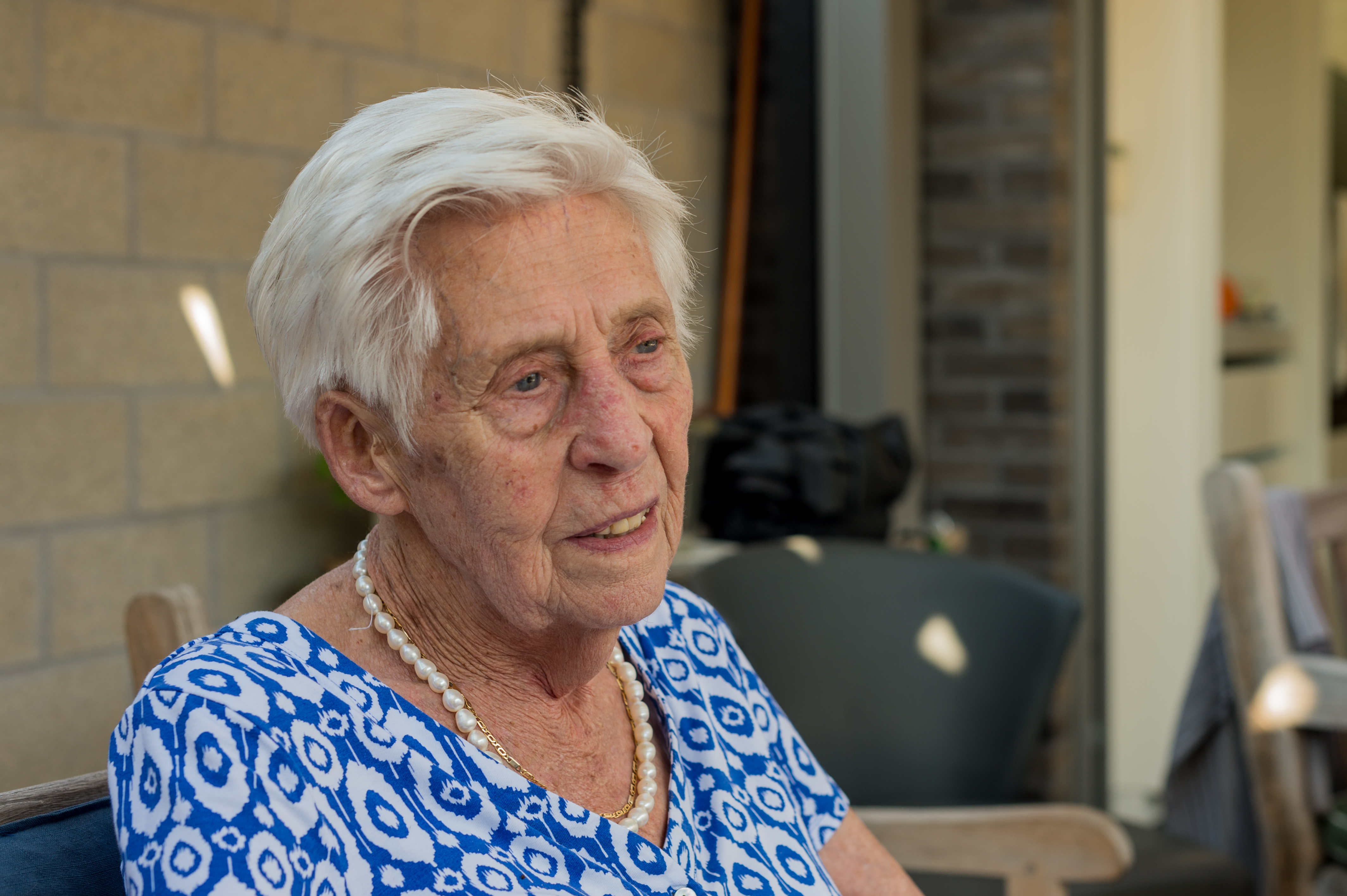 An elderly woman with short white hair sits outside, wearing a blue patterned dress and a pearl necklace.