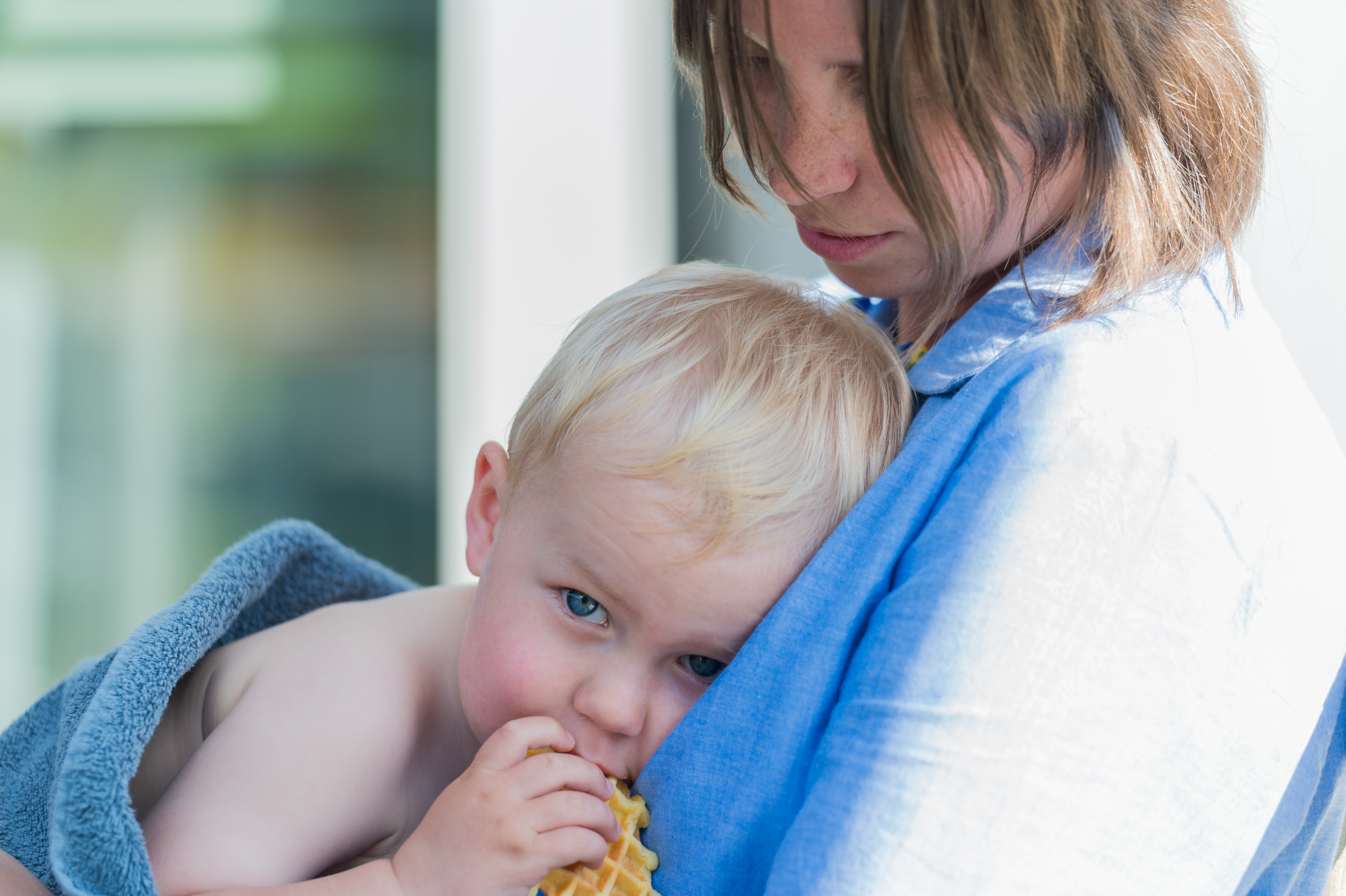 A young child wrapped in a towel eats a waffle while being held by an adult wearing a blue shirt.