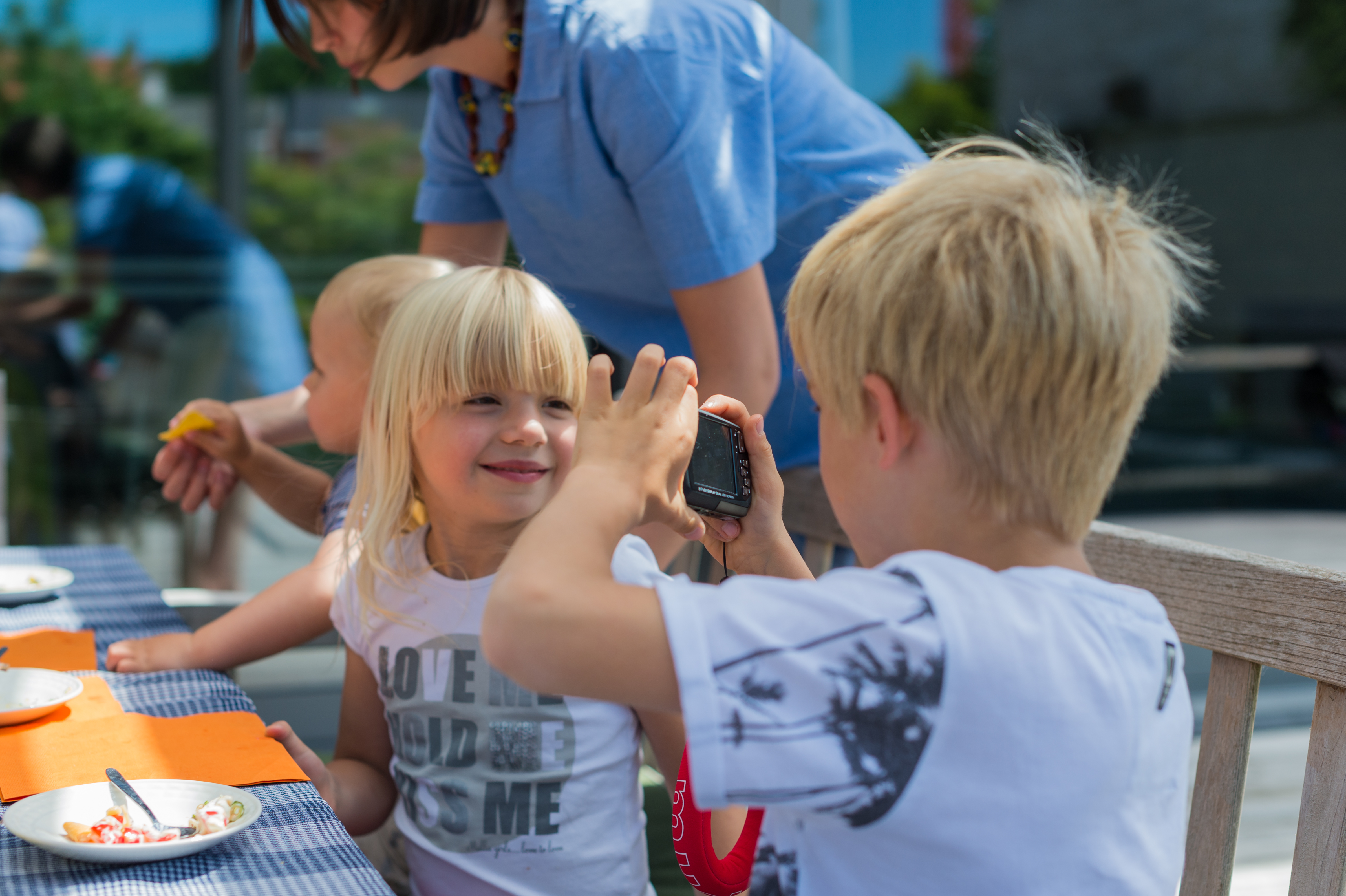 A child holds a small camera, taking a photo of a smiling girl sitting at a table during a gathering.