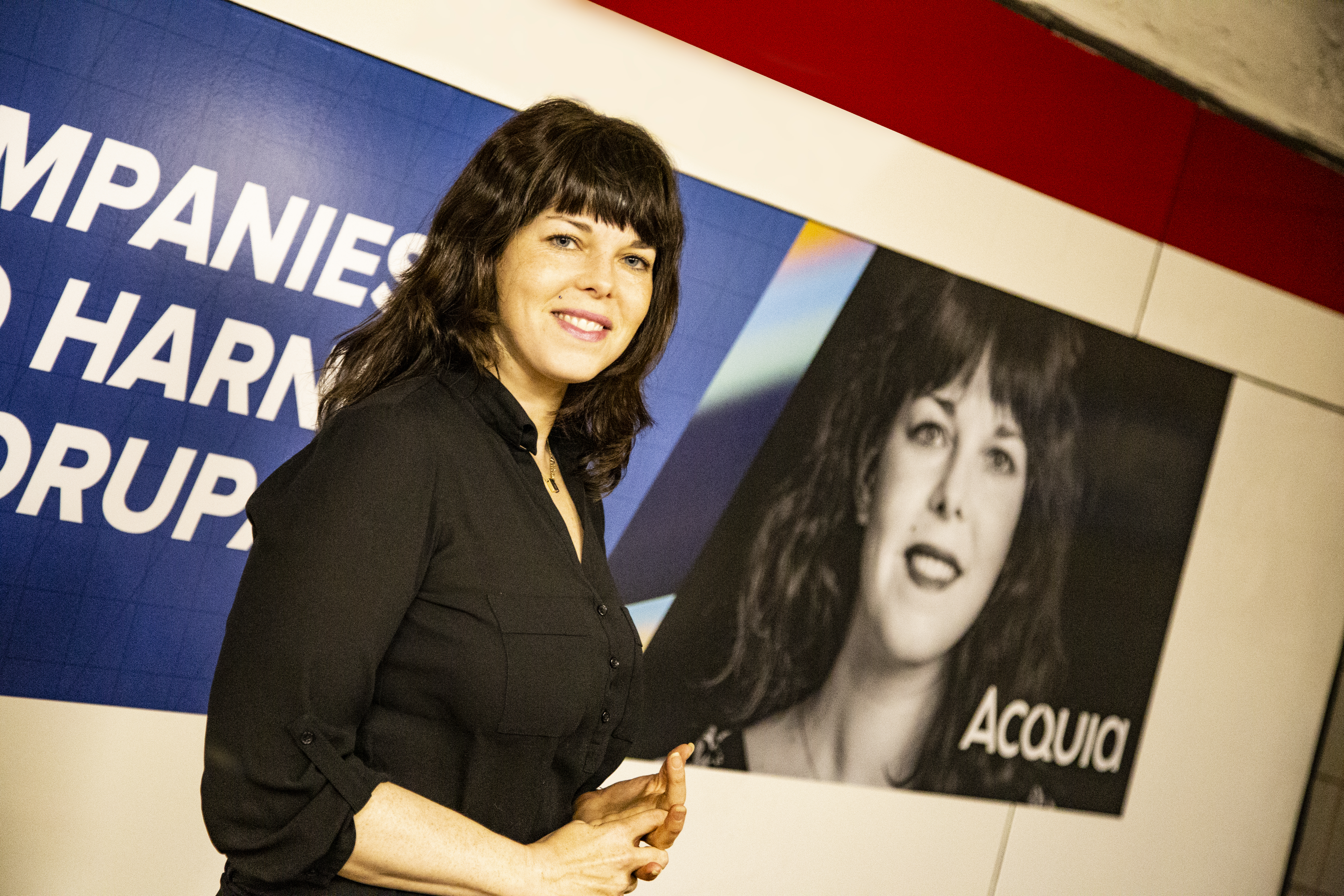 A woman in a black shirt stands in front of a wall with her photo and the word "Acquia.