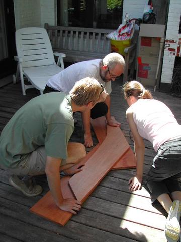 Three people kneel on a wooden deck, assembling and examining wooden boat pieces together.