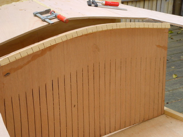 Partially built wooden boat with curved plywood panel, secured with clamps.