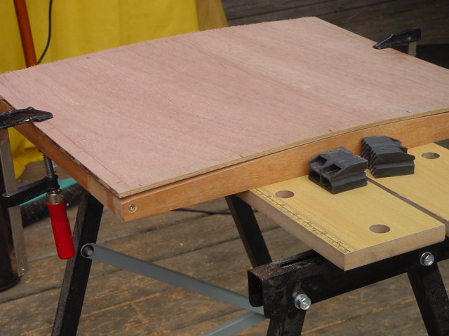 A curved wooden boat panel is clamped to a workbench, with plastic clamps resting on the surface.
