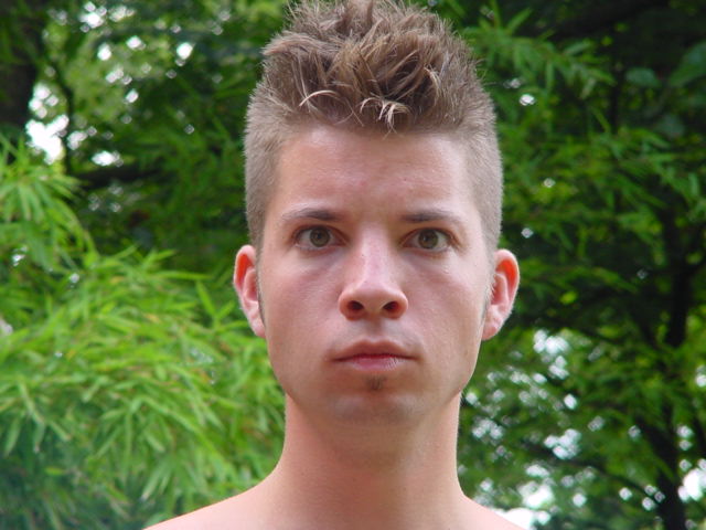 A person with short, spiky hair looks directly at the camera with a serious expression, standing outdoors.
