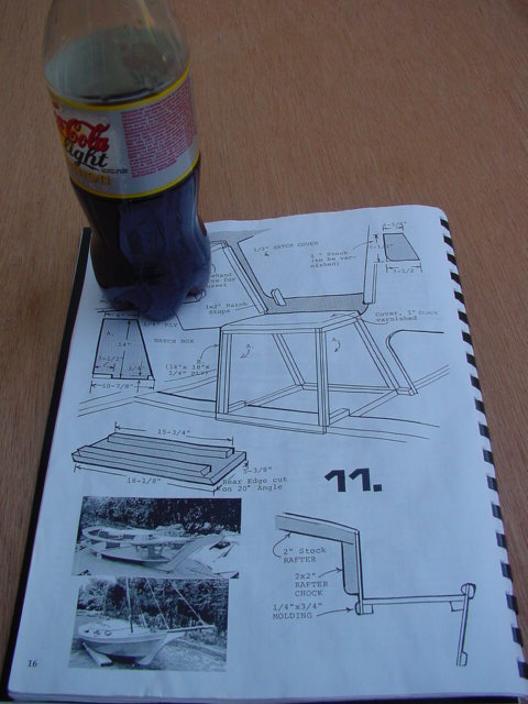 An open book with boat construction diagrams and measurements, with a plastic soda bottle resting on the page.