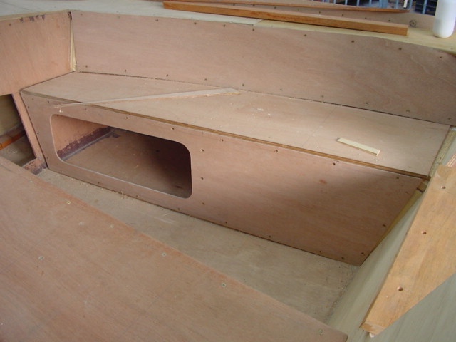 Partially built wooden boat interior with bench seating and storage compartment, showing unfinished construction with visible screws and wood pieces.