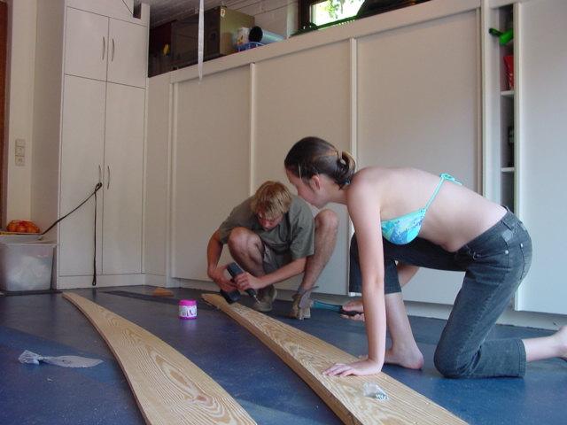 Two people kneel on the floor, working together to assemble wooden boat parts in a workshop.
