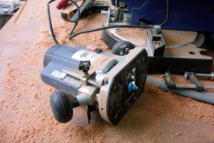 A handheld wood router rests on a workbench covered in wood shavings, with tools and clamps nearby.