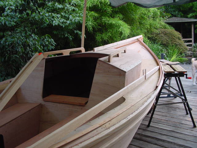 A partially built wooden boat sits on a deck, with tools and a workbench nearby.