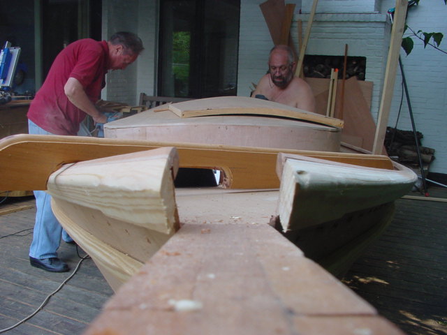 Two men work on building a wooden boat, shaping and assembling its structure on a deck.