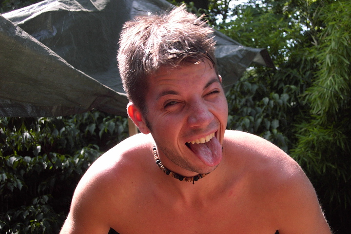 A shirtless person outdoors making a playful face, sticking out their tongue and squinting in the sunlight.