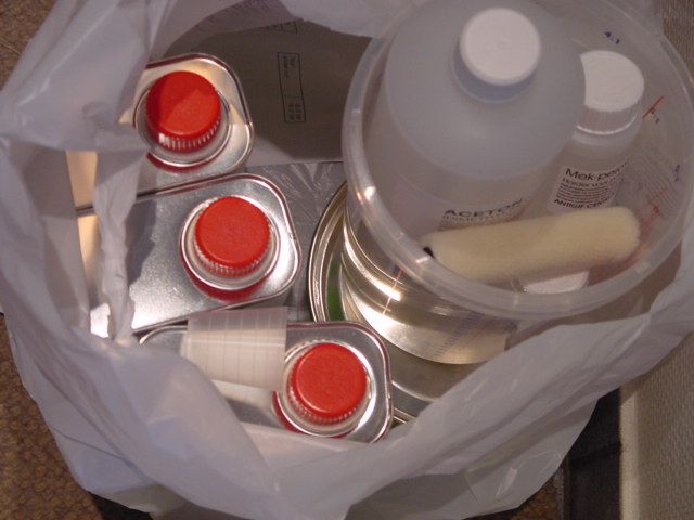 A plastic bag holds cans, bottles, and a brush, likely containing boat-building supplies like solvents or adhesives.