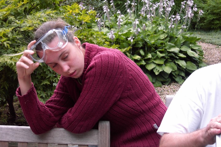 A person in a red sweater with safety goggles rests their head on a bench, appearing tired.