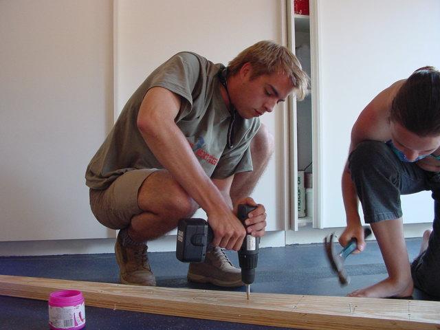 A man drills into a wooden plank while a woman hammers nearby, both working on a boat-building project.